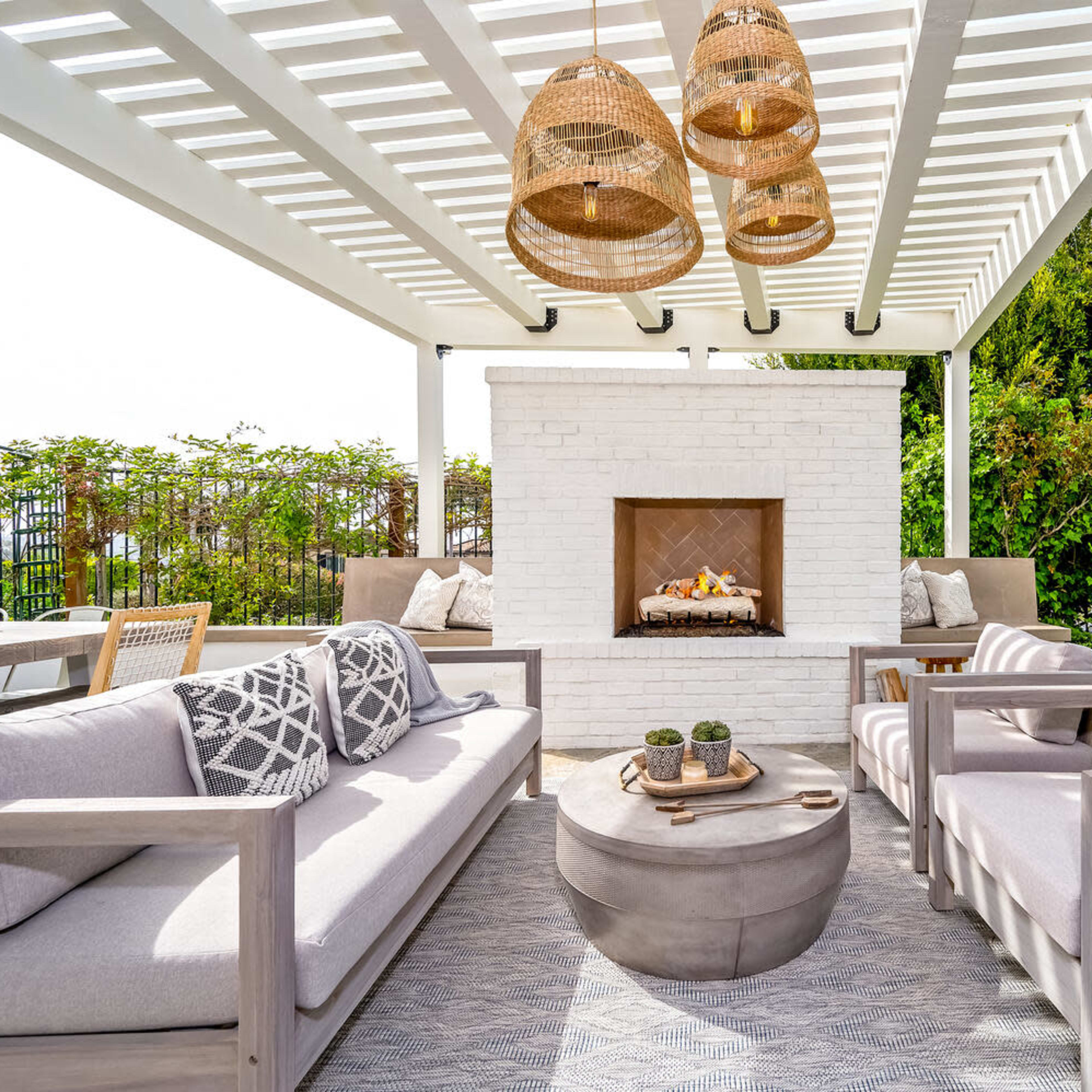 A stylish outdoor lounge area with two gray sofas, a round coffee table, and a white brick fireplace, all set under a white pergola with hanging light fixtures.