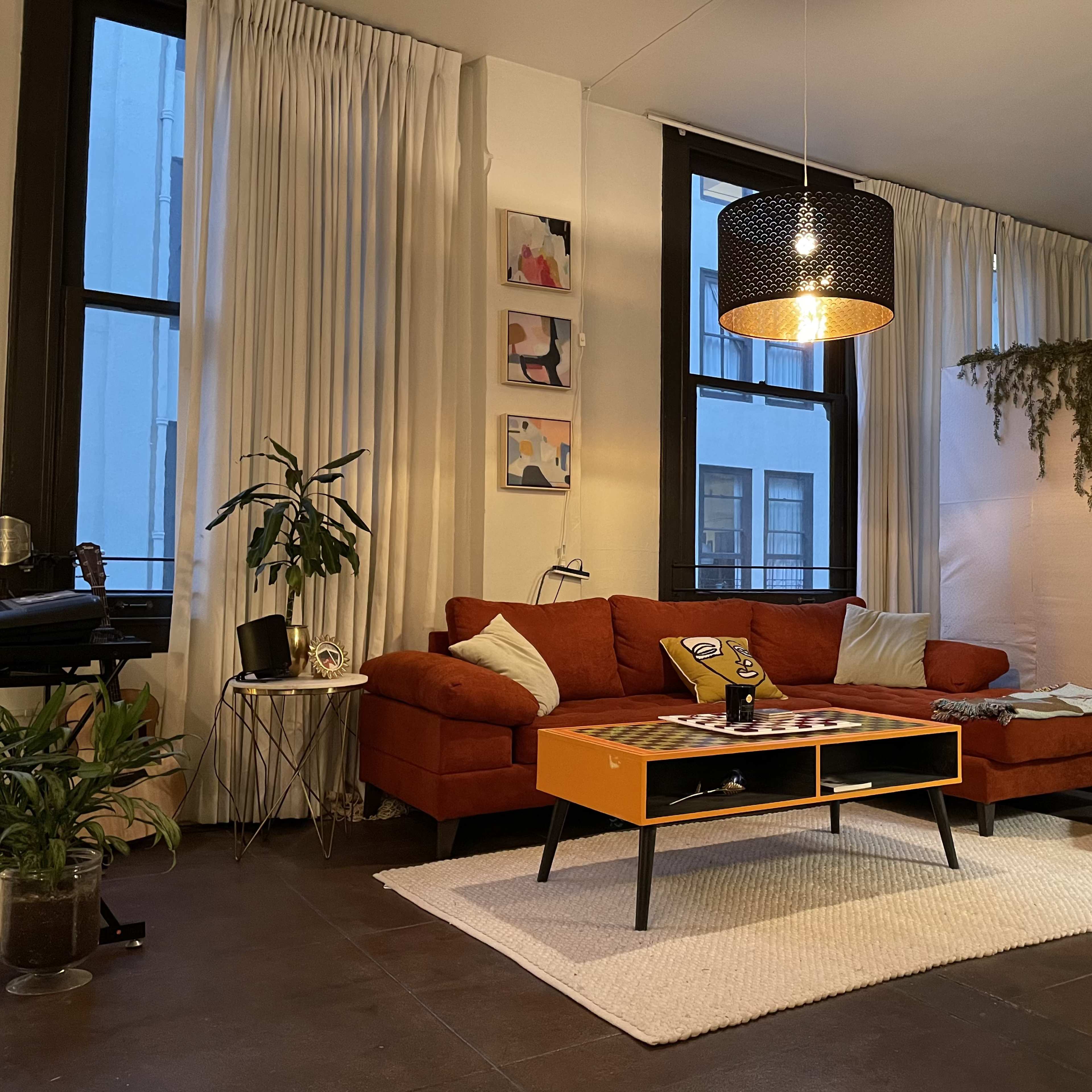 A cozy living room featuring a brown sectional sofa, an orange coffee table, large windows with sheer curtains, and various plants throughout the space.