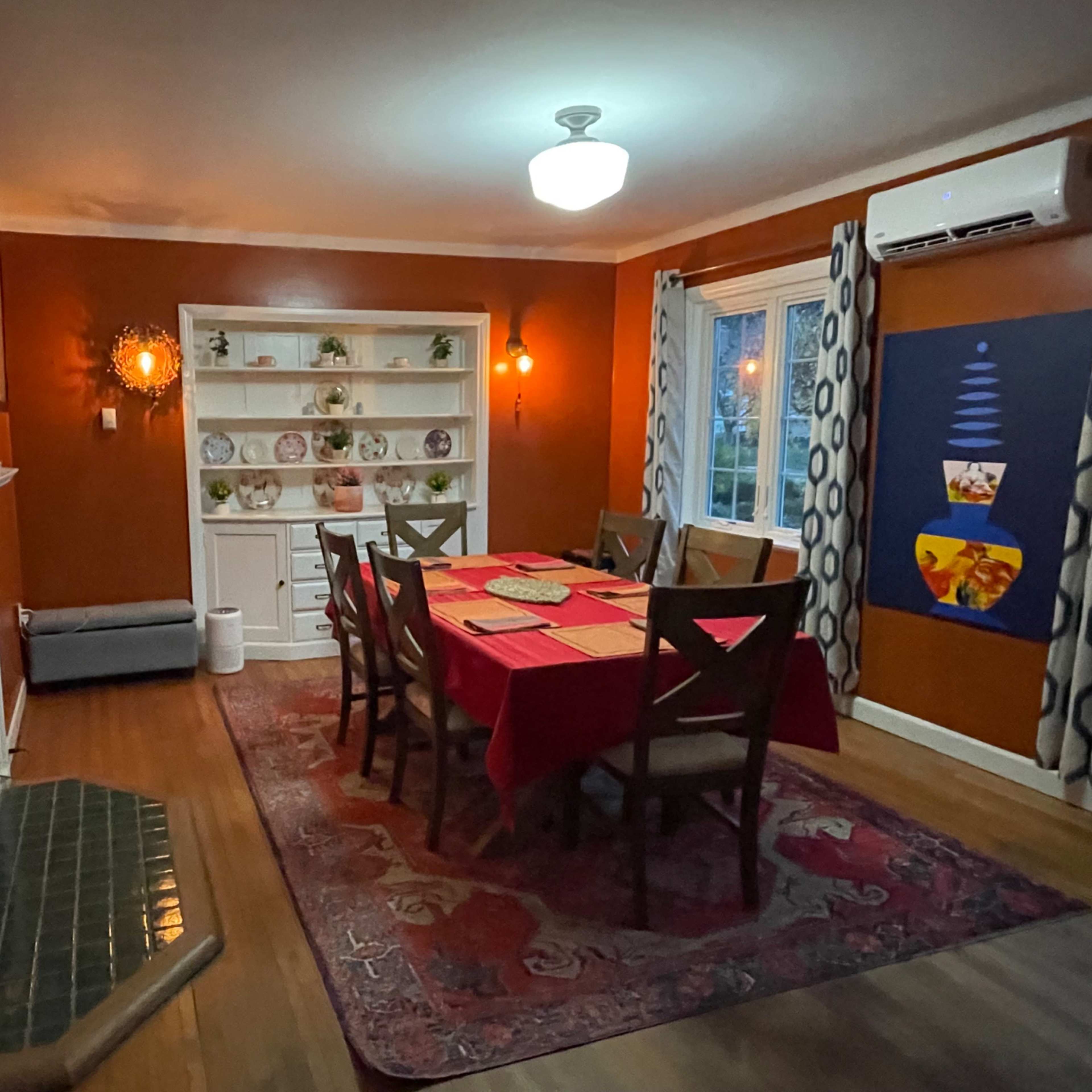 A dining room features a red tablecloth over a large table surrounded by chairs, with a fireplace and shelves displaying dishes along the walls.