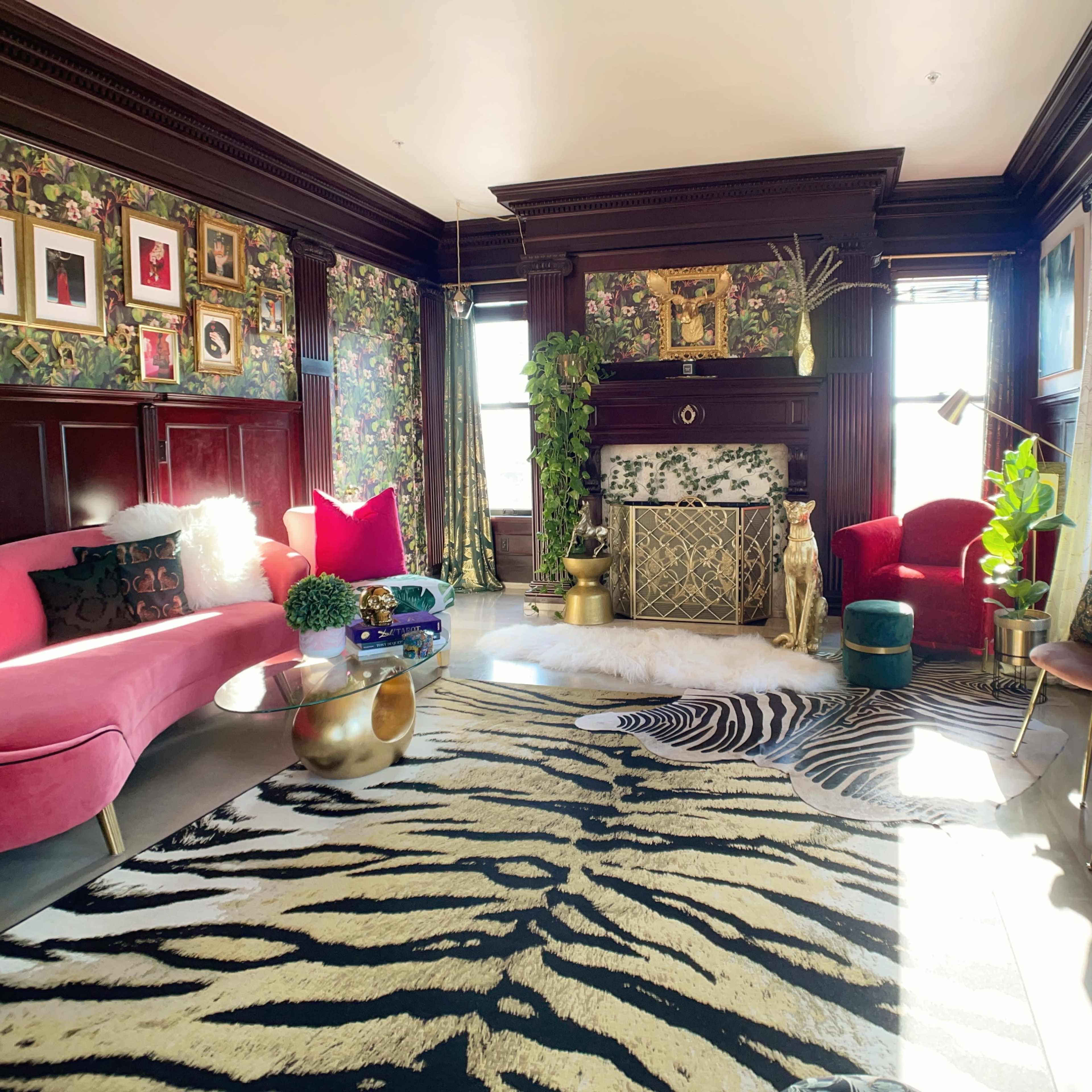 A vibrant living room with a pink sofa, red armchairs, a patterned rug, and various decorative plants and artwork.