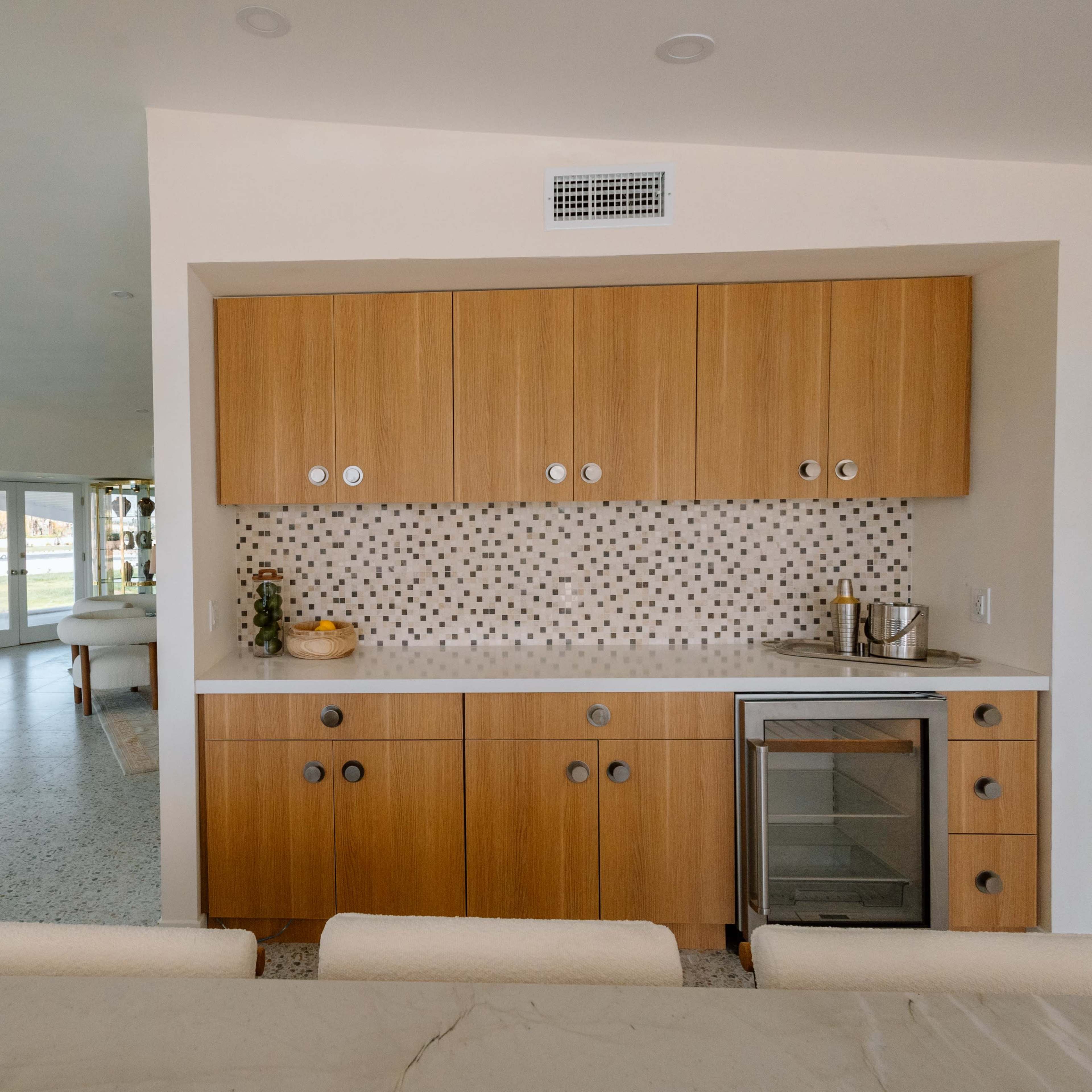 A modern kitchen with wooden cabinets, a white countertop, and a wine cooler, adjacent to a spacious living area with large windows.