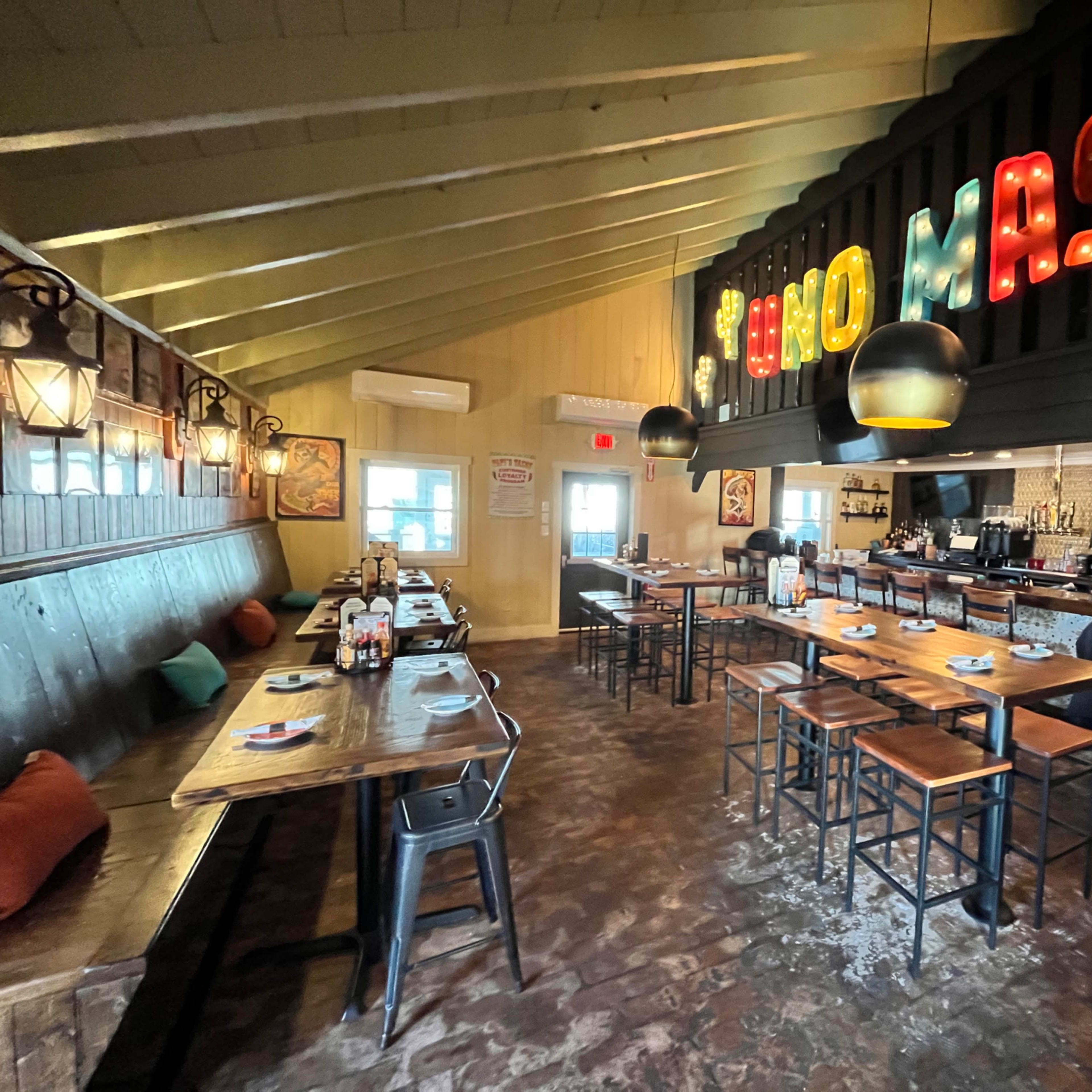 An interior of a restaurant with wooden tables, chairs, and a bar area illuminated by colorful lights.