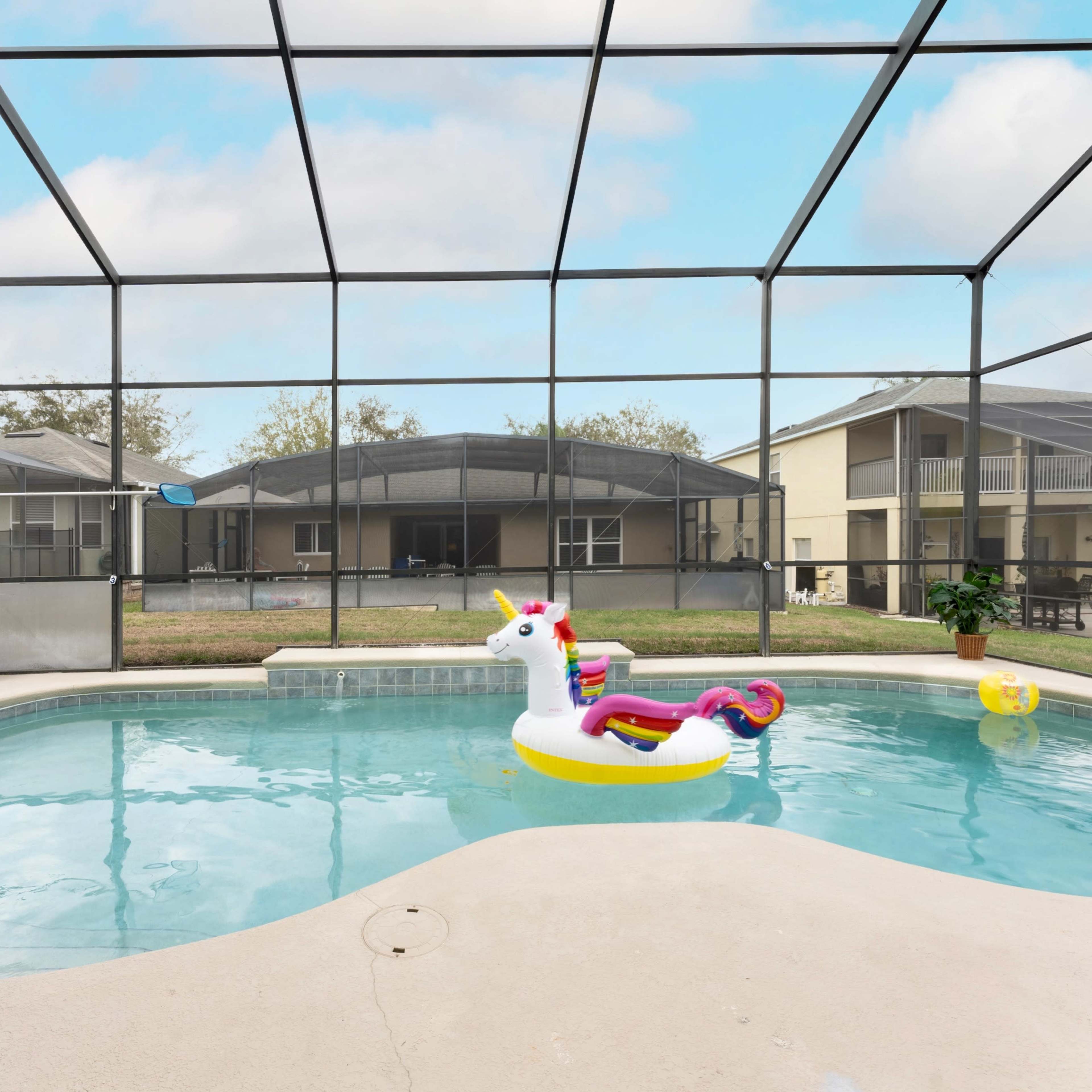 A colorful inflatable unicorn floats in a private pool, surrounded by a screened enclosure and residential buildings in the background.