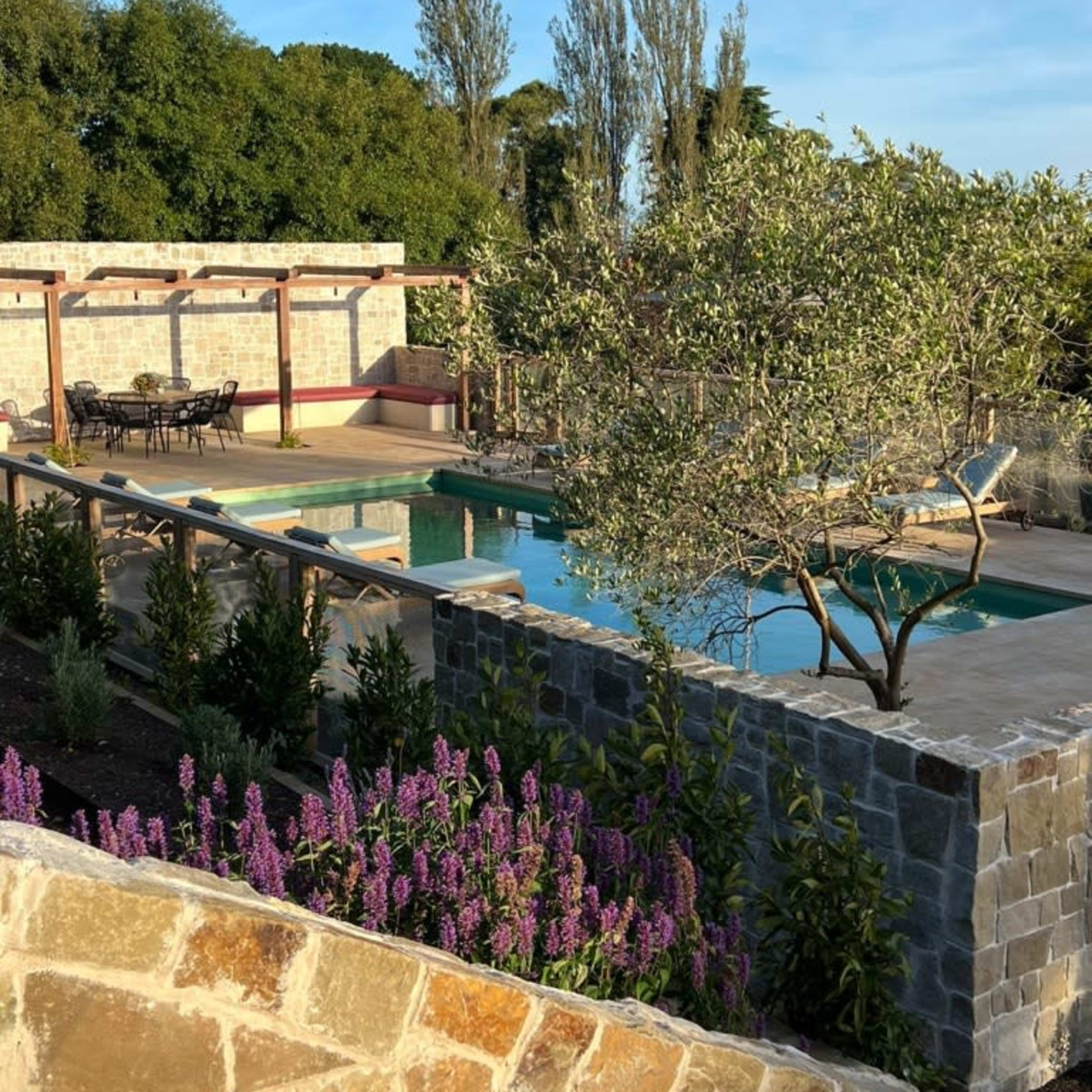 A landscaped area featuring a swimming pool surrounded by stone walls and vibrant purple flowers, with outdoor seating visible in the background.