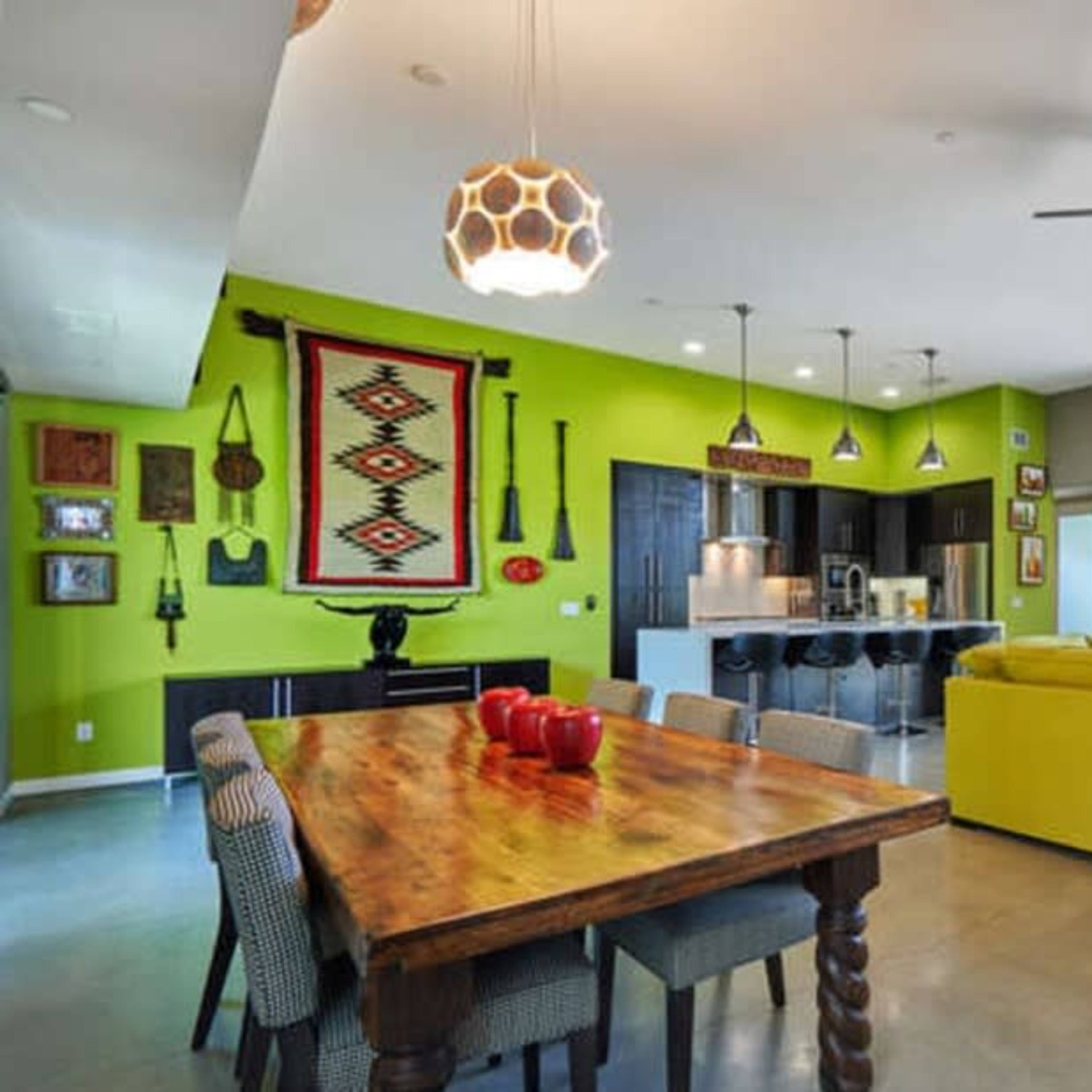 A modern living space with a green accent wall, a wooden dining table, a bright yellow sofa, and large windows overlooking a patio and pool area.