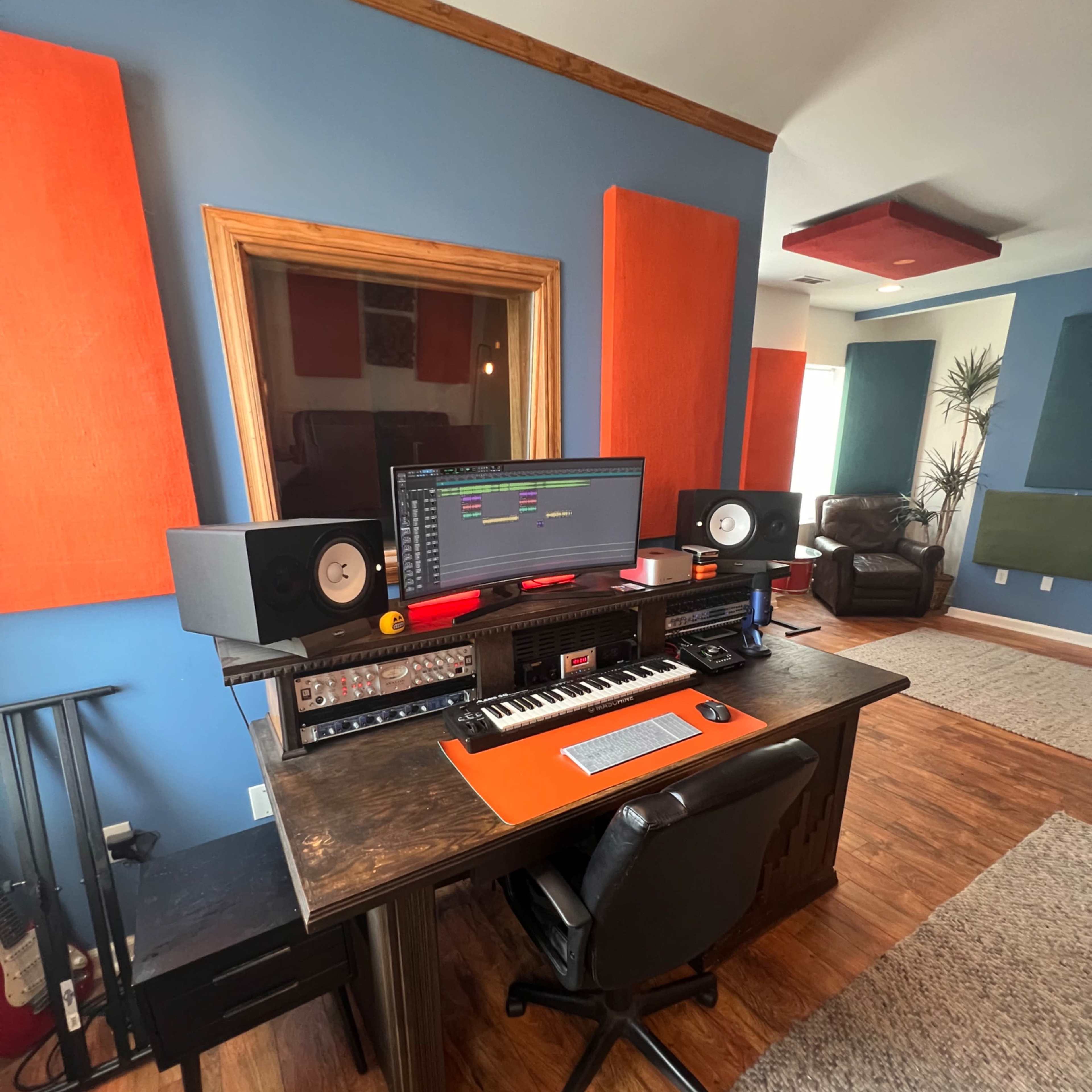 A music production studio features a desk with a computer monitor, speakers, and a keyboard, surrounded by acoustic panels and comfortable seating.