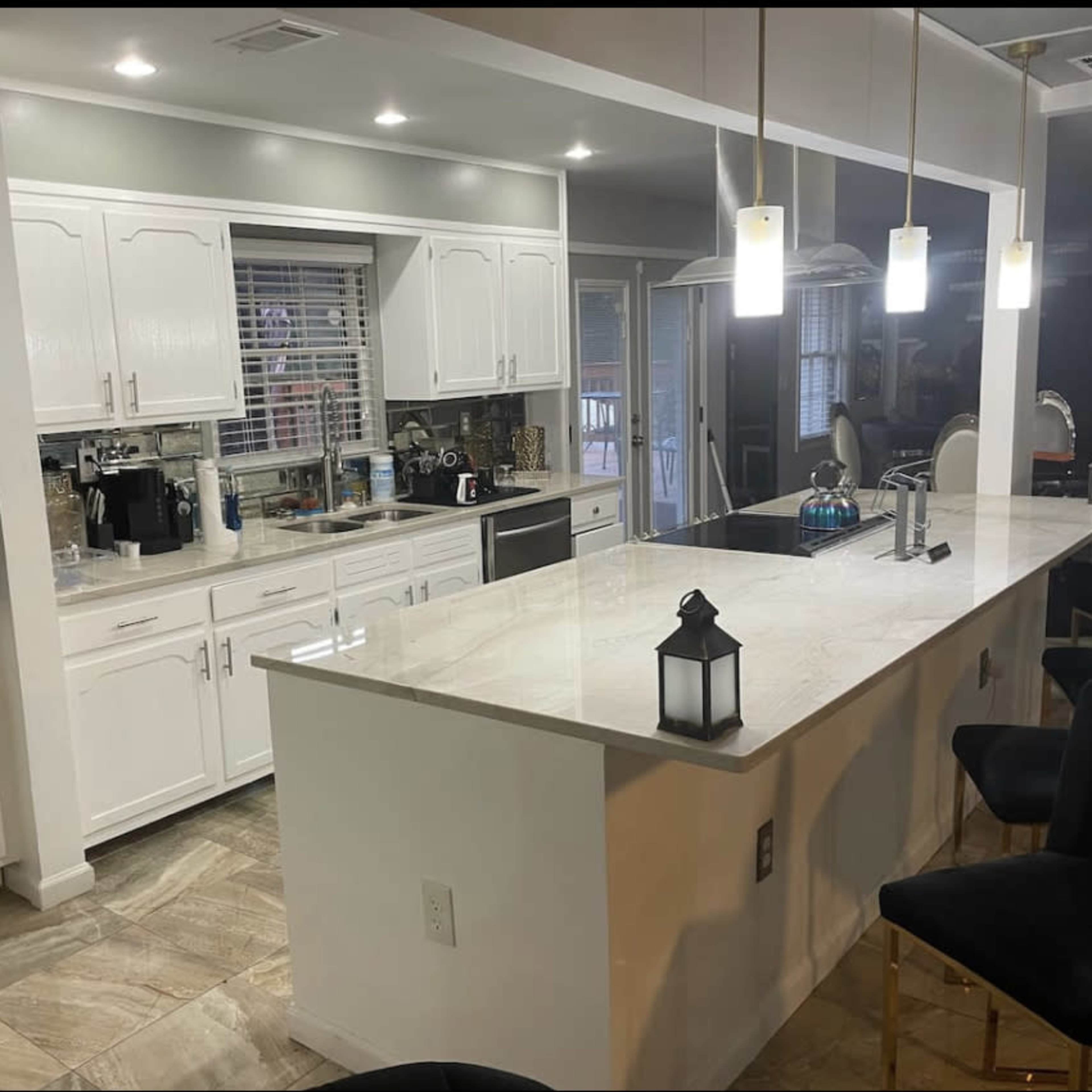 A modern kitchen with white cabinetry, a central island with bar seating, and pendant lights, featuring stainless steel appliances and a window overlooking an outdoor area.