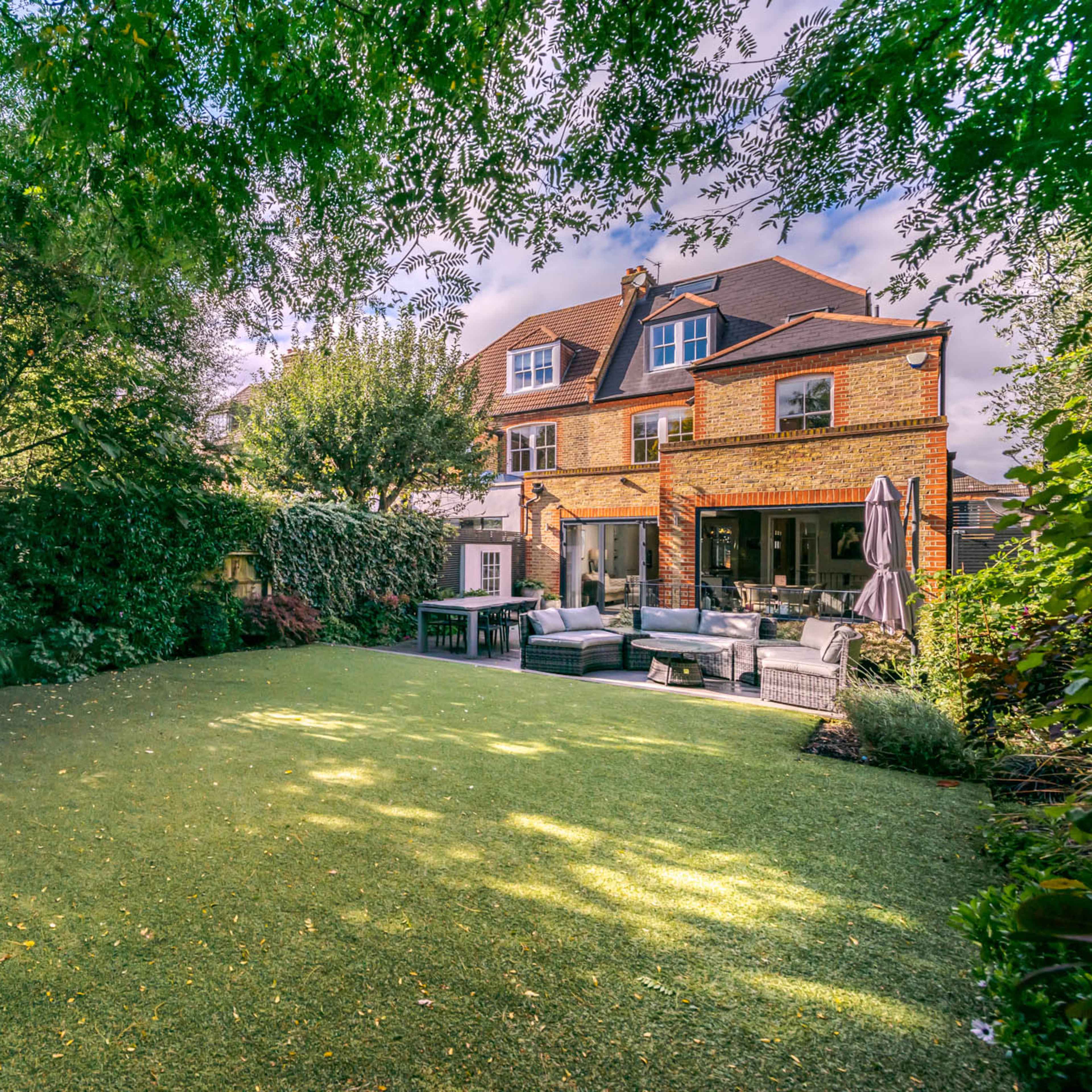 A well-maintained backyard with artificial grass, surrounded by lush greenery and featuring a two-story brick house.