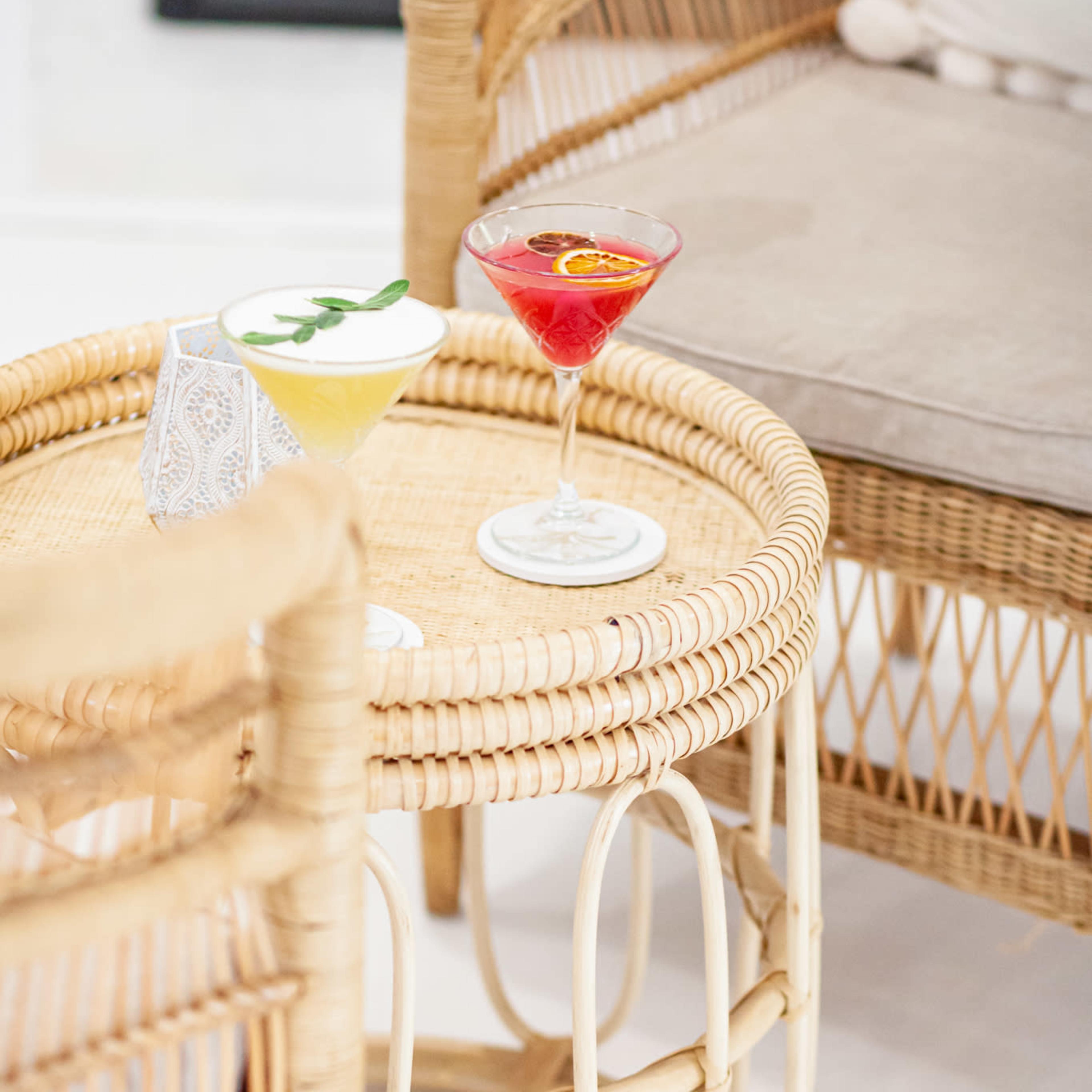 Two cocktails are placed on a woven rattan table beside a chair in a modern, light-colored interior setting.