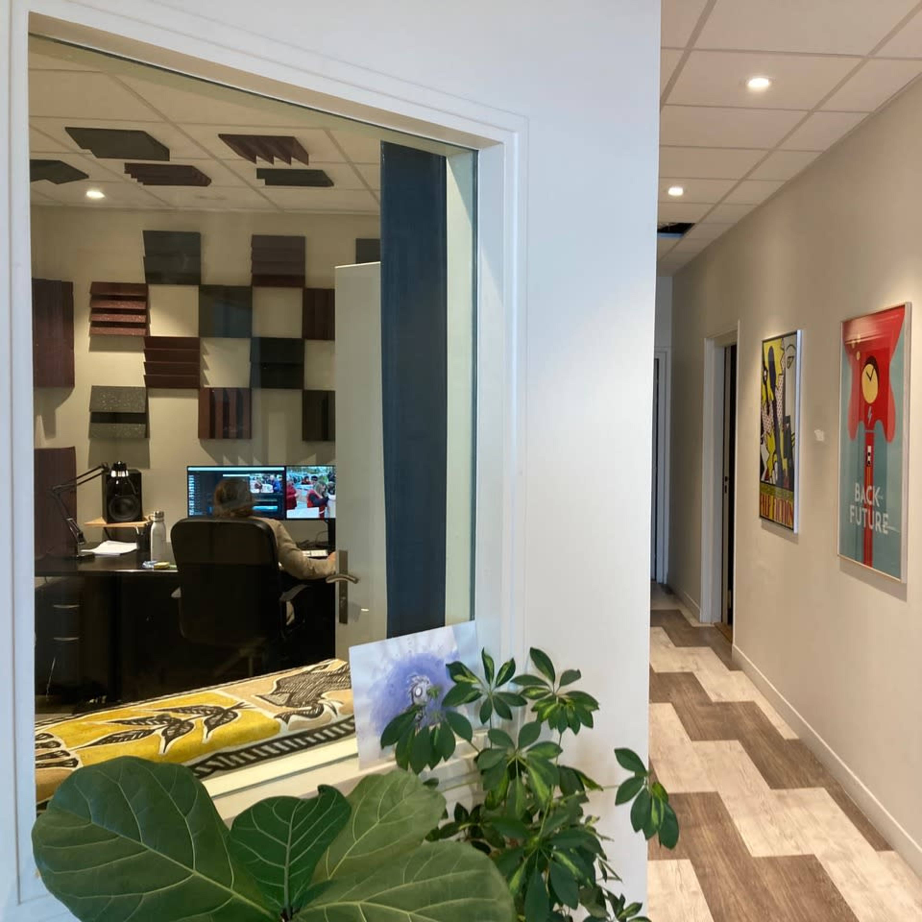 A hallway with framed posters on the walls and a window into a room with a computer setup behind it.