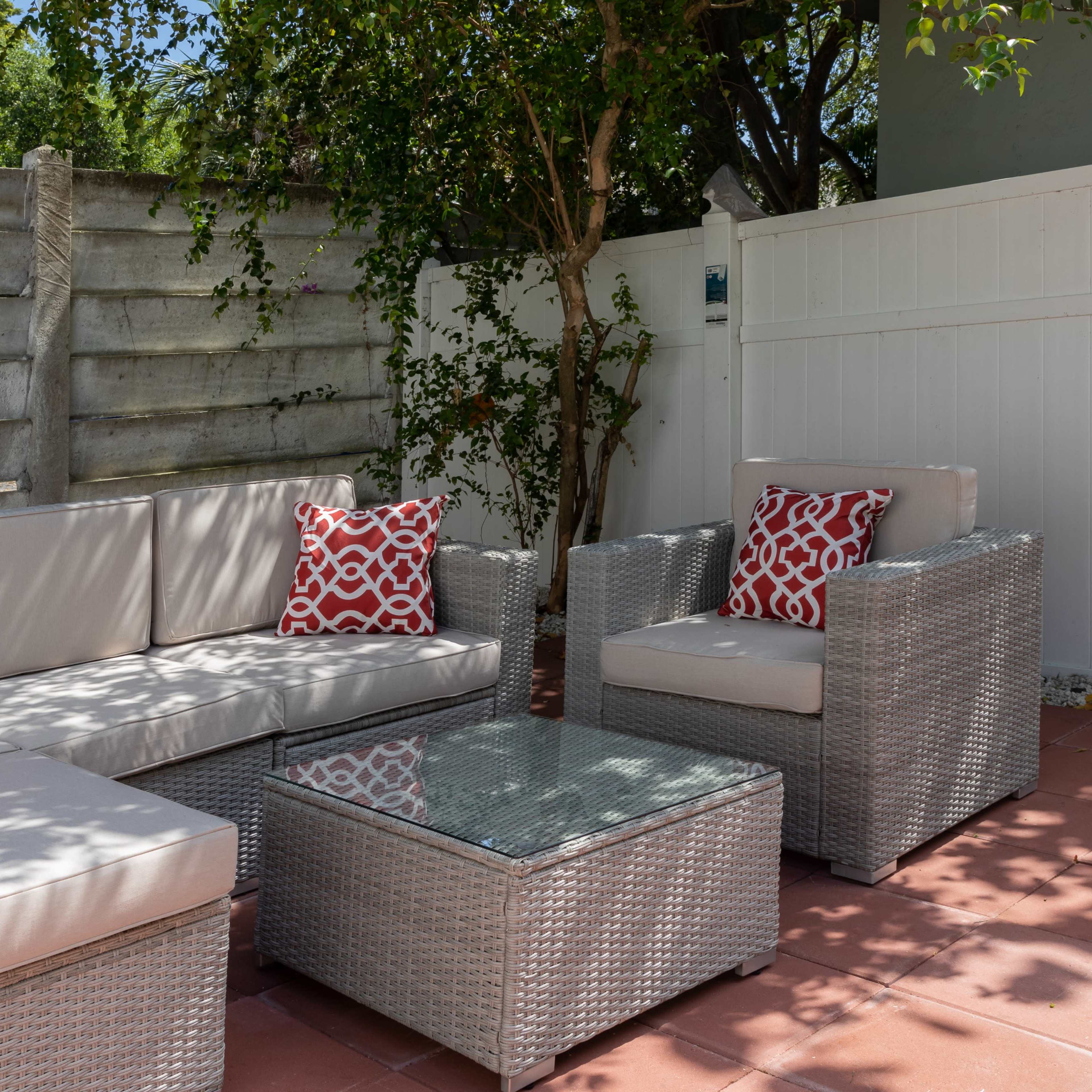 A modern outdoor seating area features a sectional sofa with red-patterned cushions, a glass-top coffee table, and a single armchair, surrounded by a concrete and wooden fence.