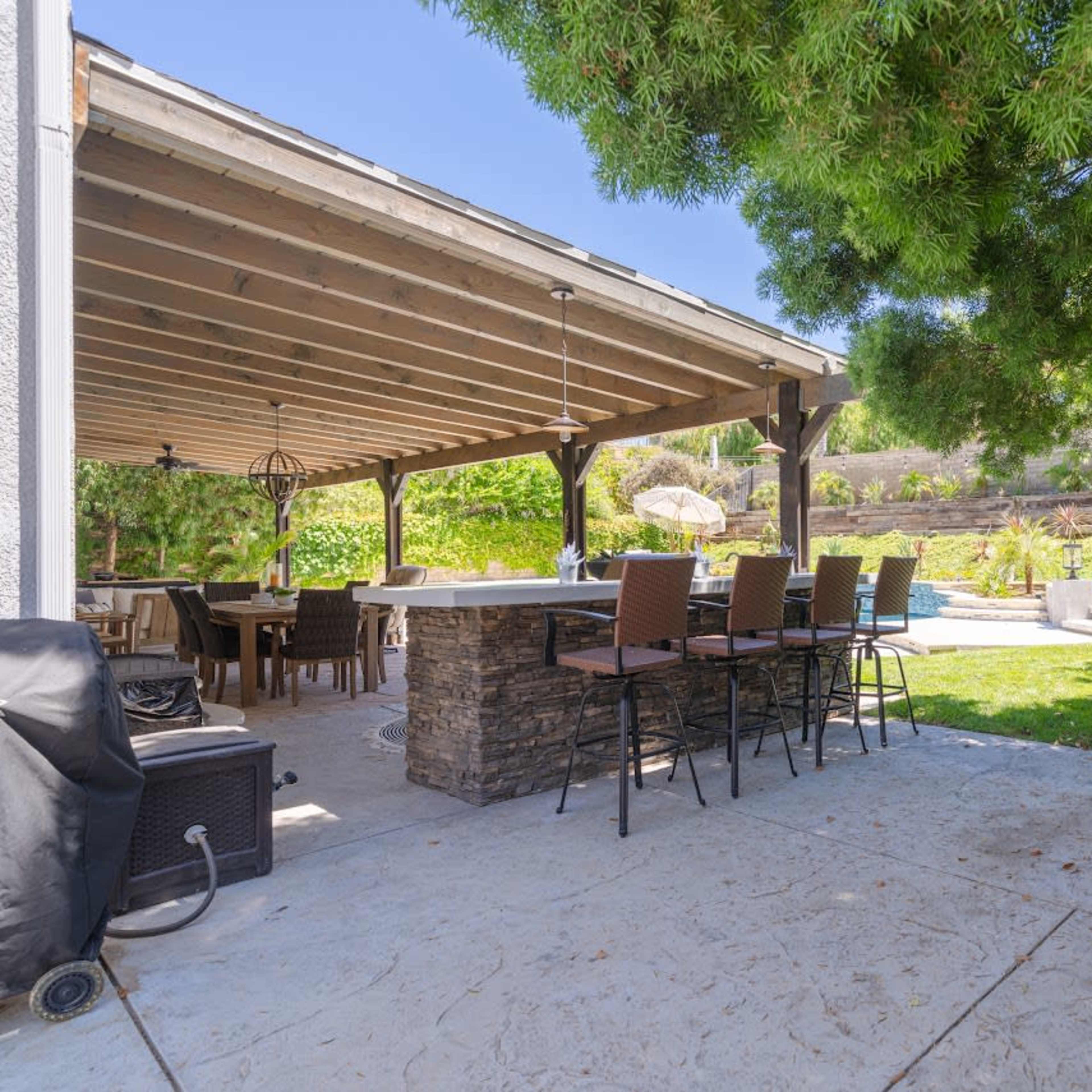 A covered patio features a bar area with stools overlooking a landscaped backyard and pool.