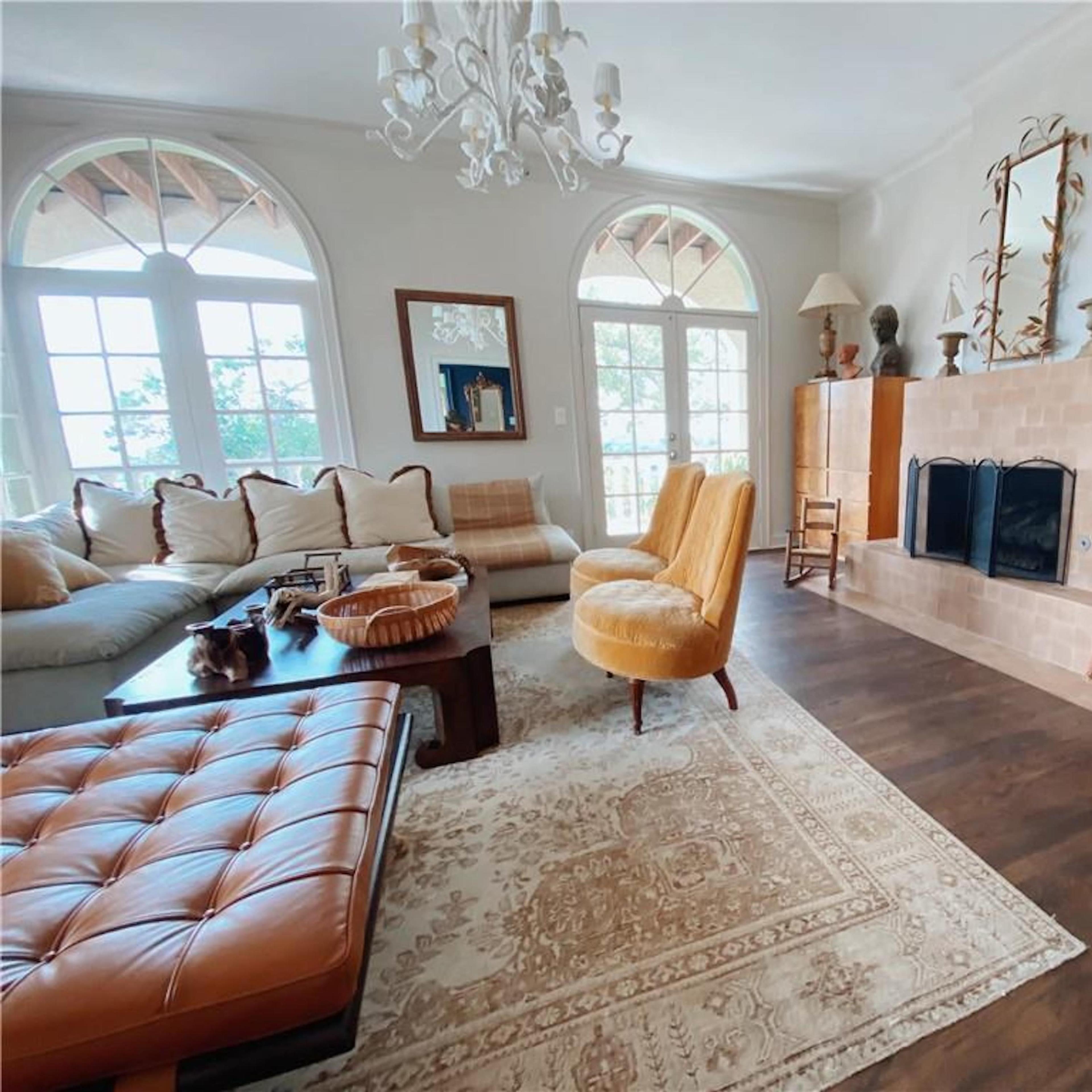 A living room with a beige sofa, two yellow armchairs, a wooden coffee table, and a fireplace, all under a chandelier and large windows.