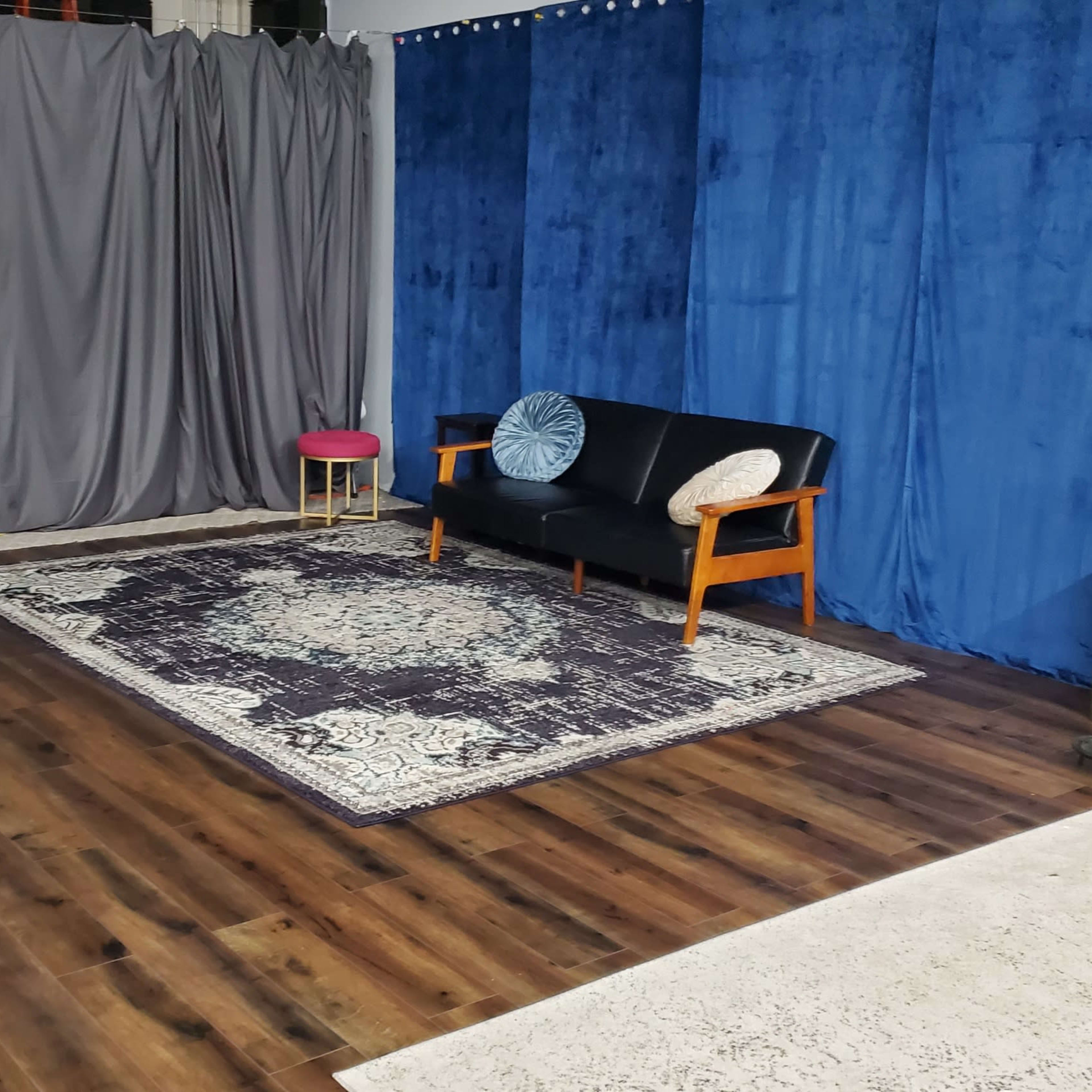 A cozy interior setup with a black sofa, a small pink stool, a patterned rug, and blue and gray fabric backdrops.