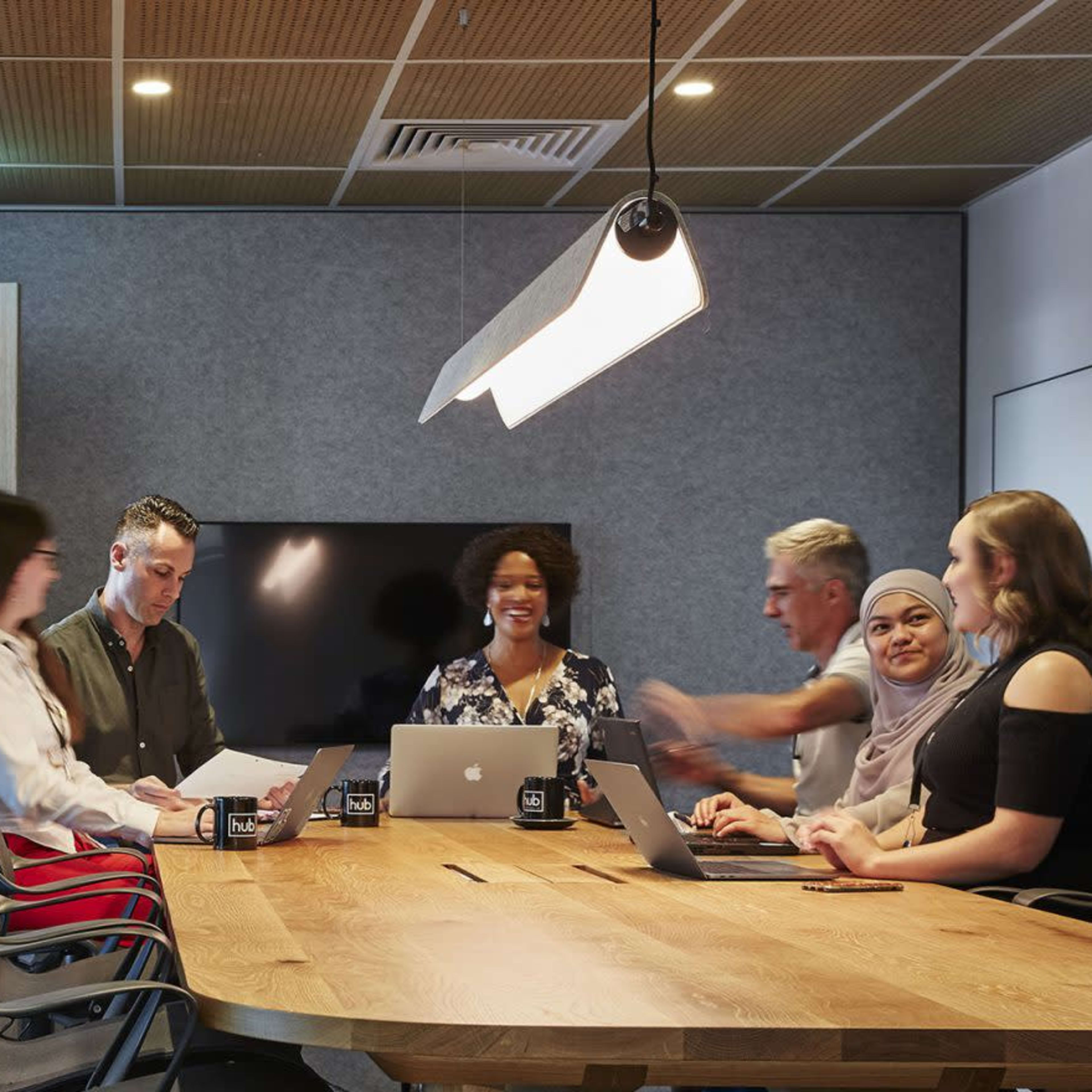 A group of seven people are sitting around a large wooden table in a modern meeting room, engaged in discussion while using laptops and papers.
