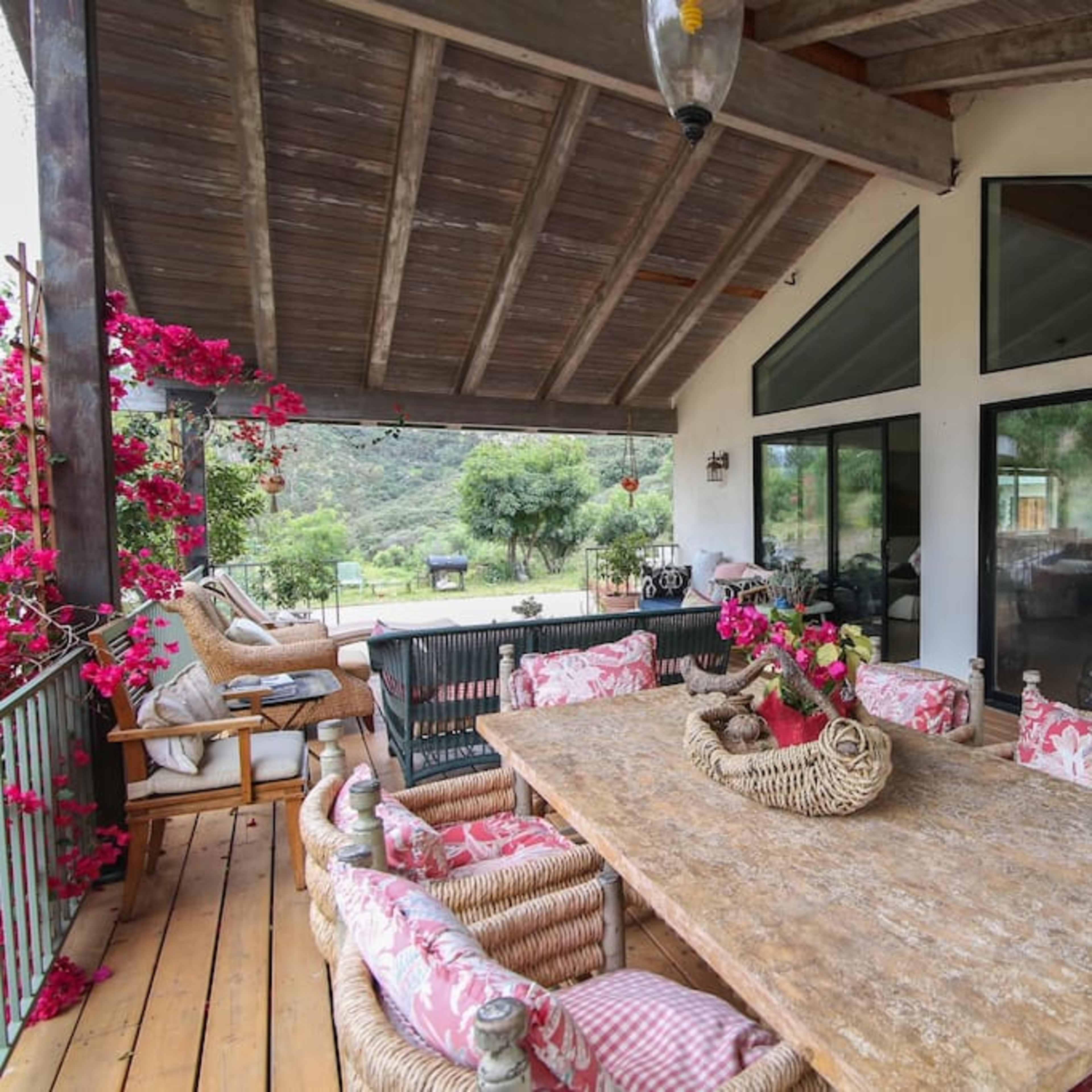 A covered patio features a large wooden table surrounded by wicker chairs, with vibrant flowers along the edges and large windows looking out onto a scenic landscape.