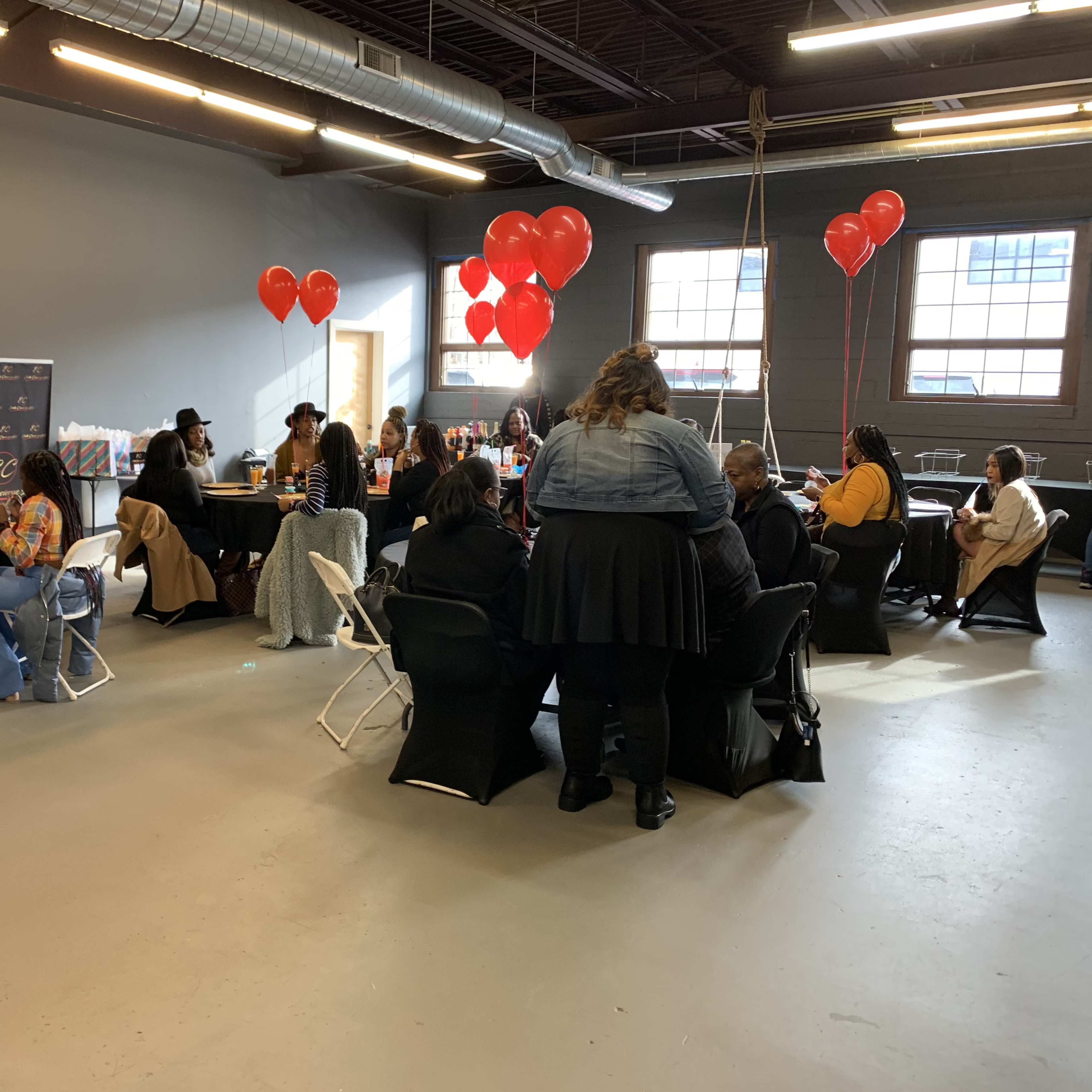 A group of people gathers inside a spacious room decorated with red balloons, seated around tables set up for an event.