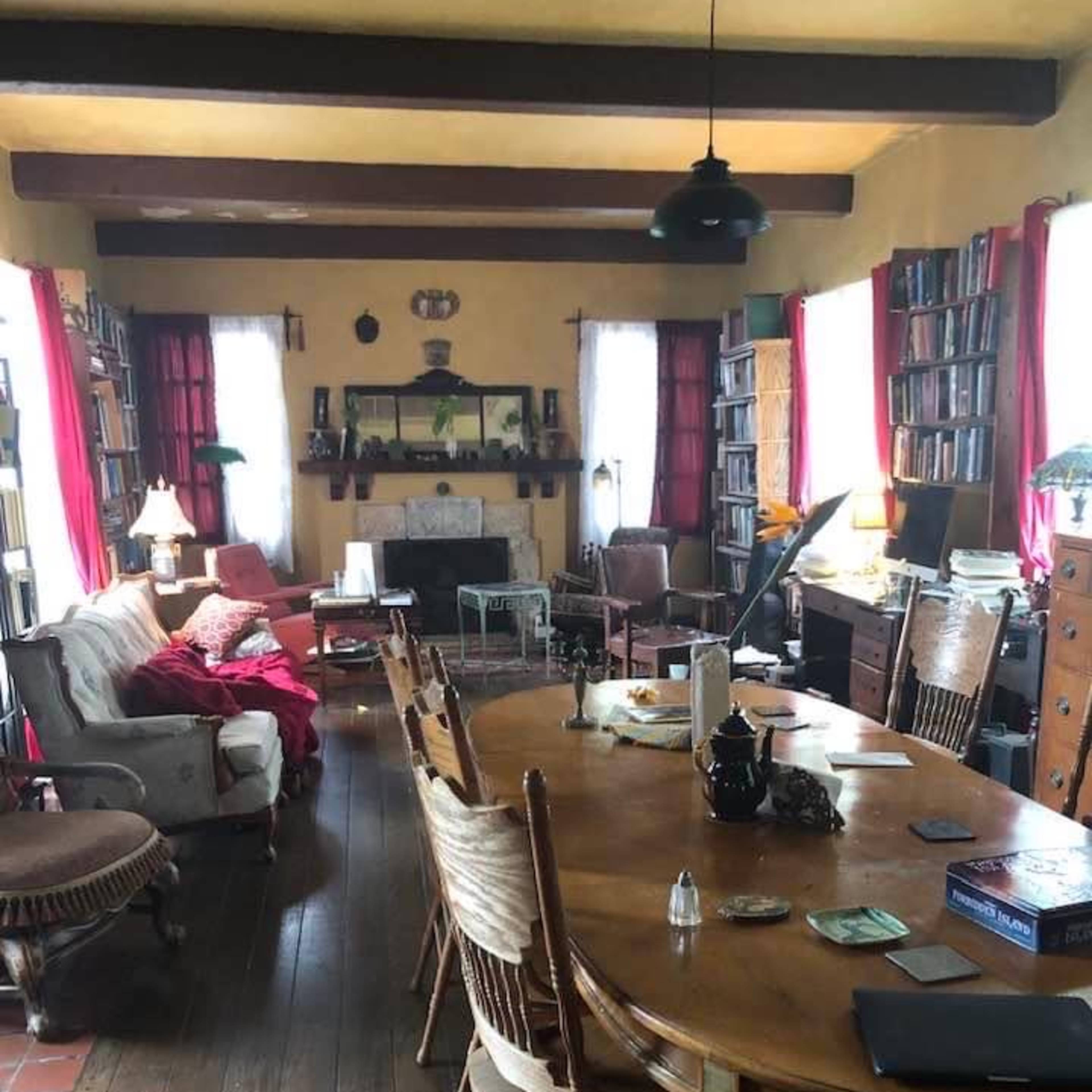 A spacious living room with a large wooden dining table, surrounded by chairs, bookshelves lining the walls, and a mix of furniture in a cozy arrangement.