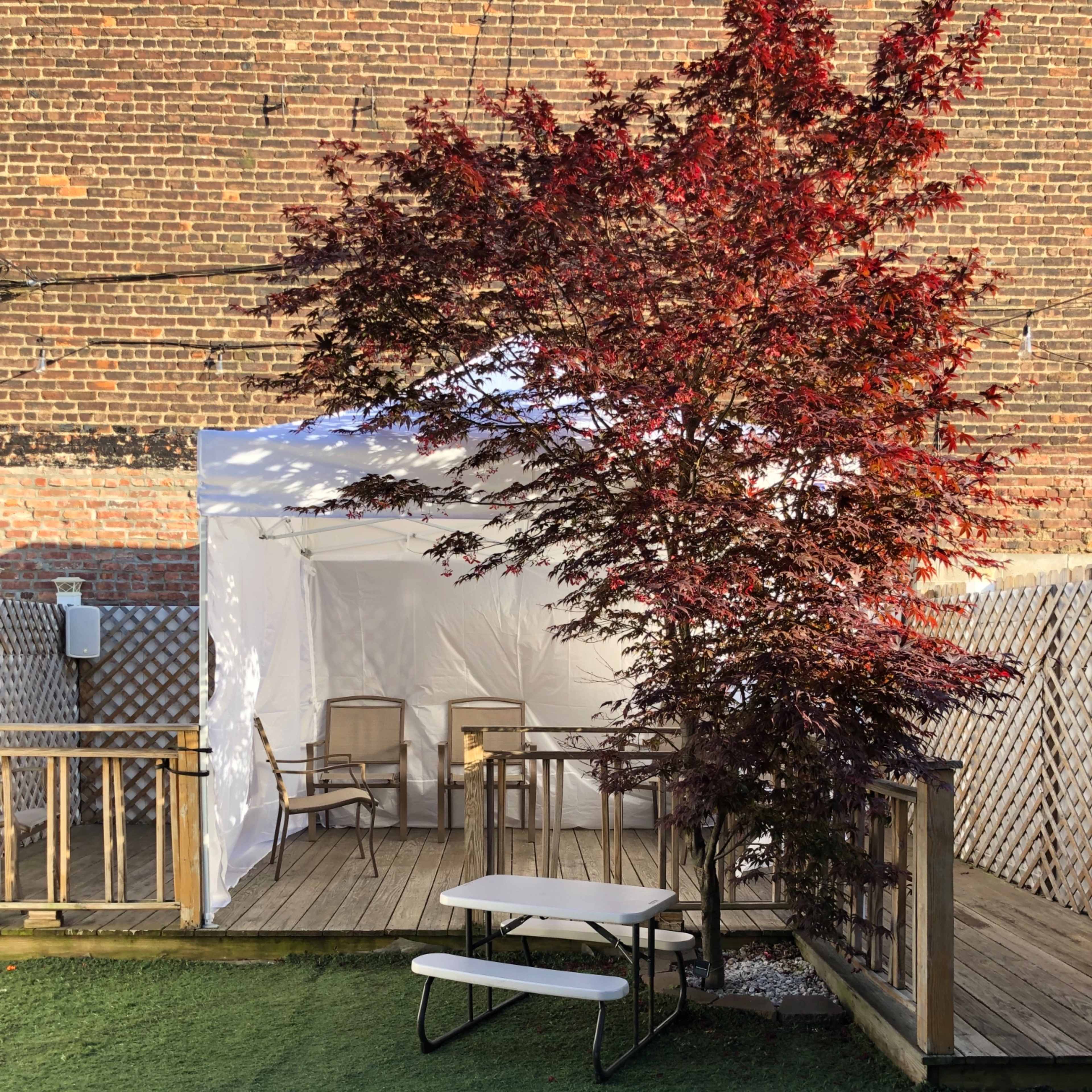 A small outdoor area features a white tent, wooden deck, a table, and chairs, surrounded by a vibrant red tree and artificial grass.