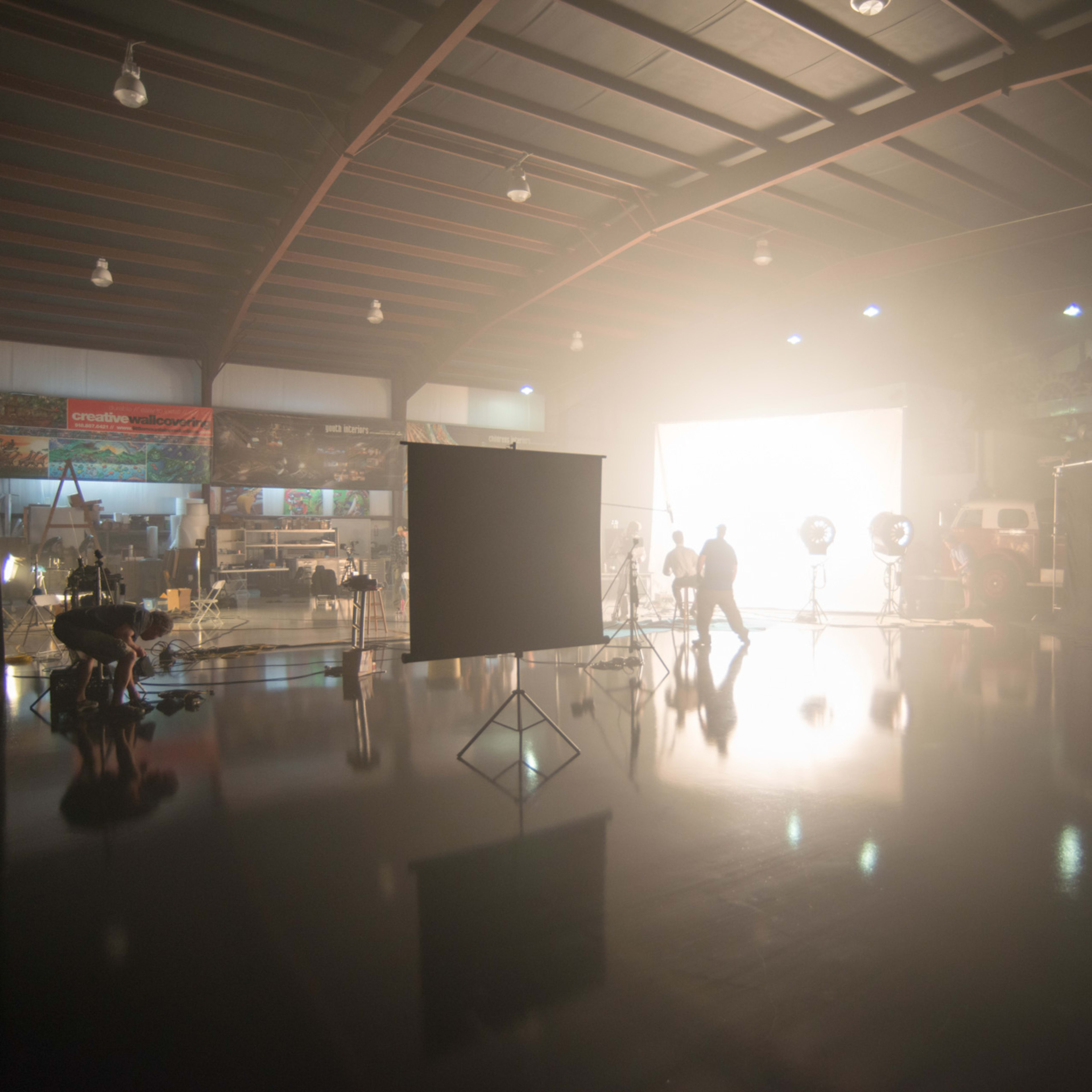 A large film studio is set up with bright lights, reflective surfaces, and various filming equipment.