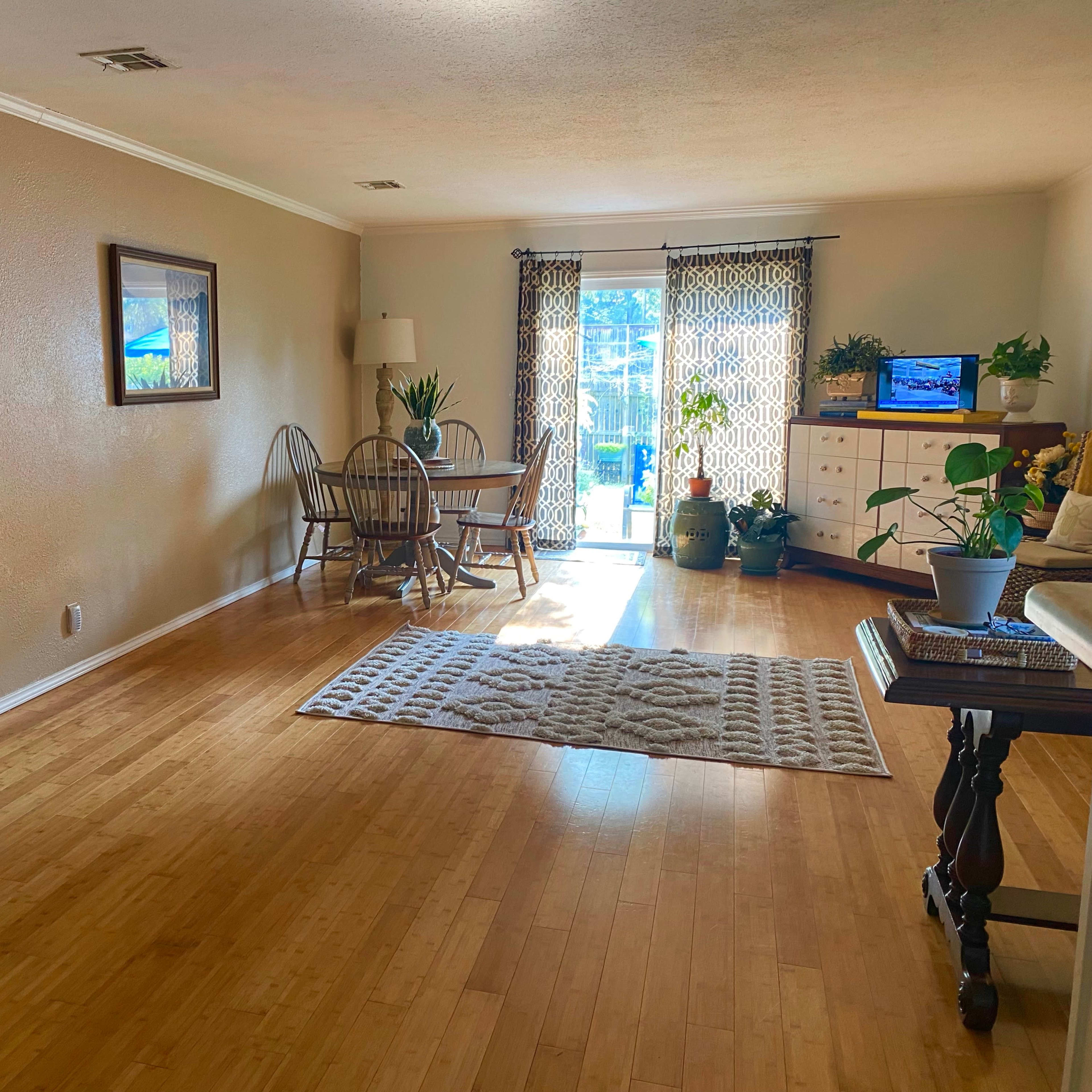The room features bamboo flooring, a dining area with a table and chairs, a small seating area with a lamp and plants, and large windows leading to a patio.