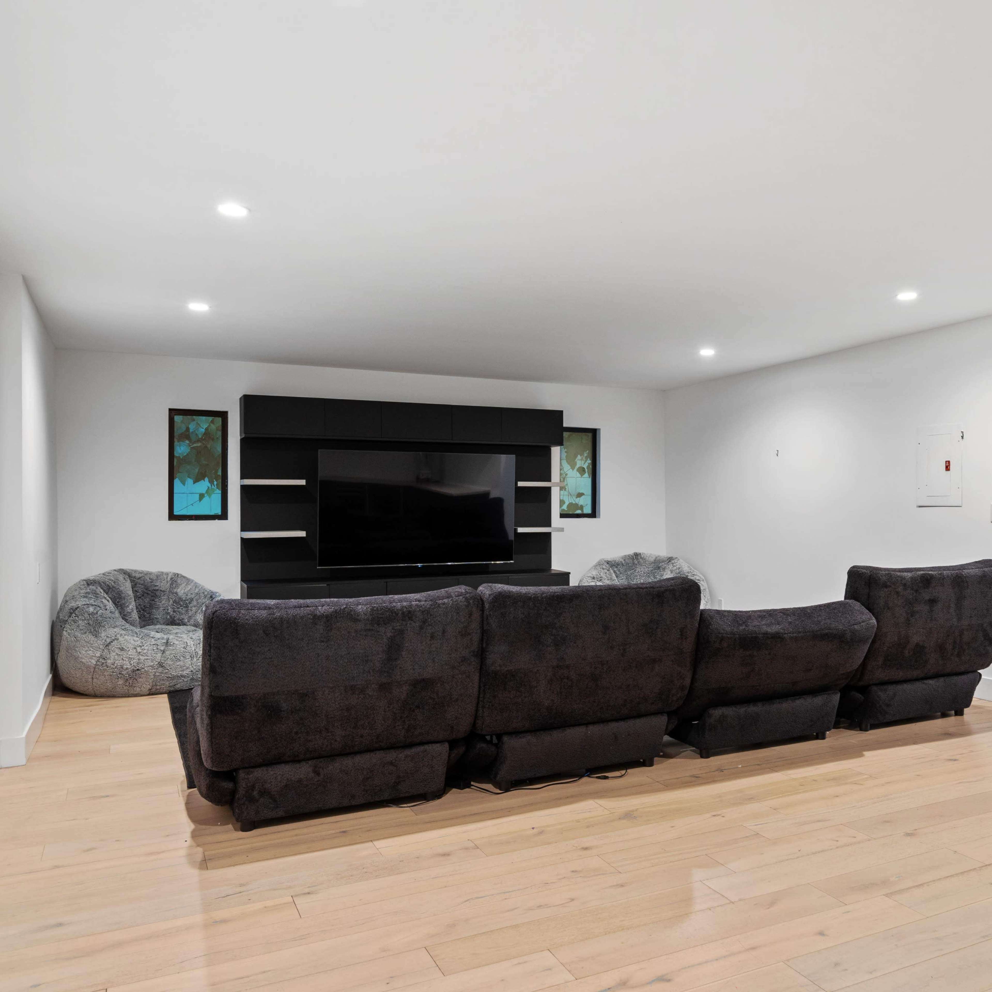 A modern living room with a black sectional sofa facing a large flat-screen TV on a wall, alongside two beanbag chairs.