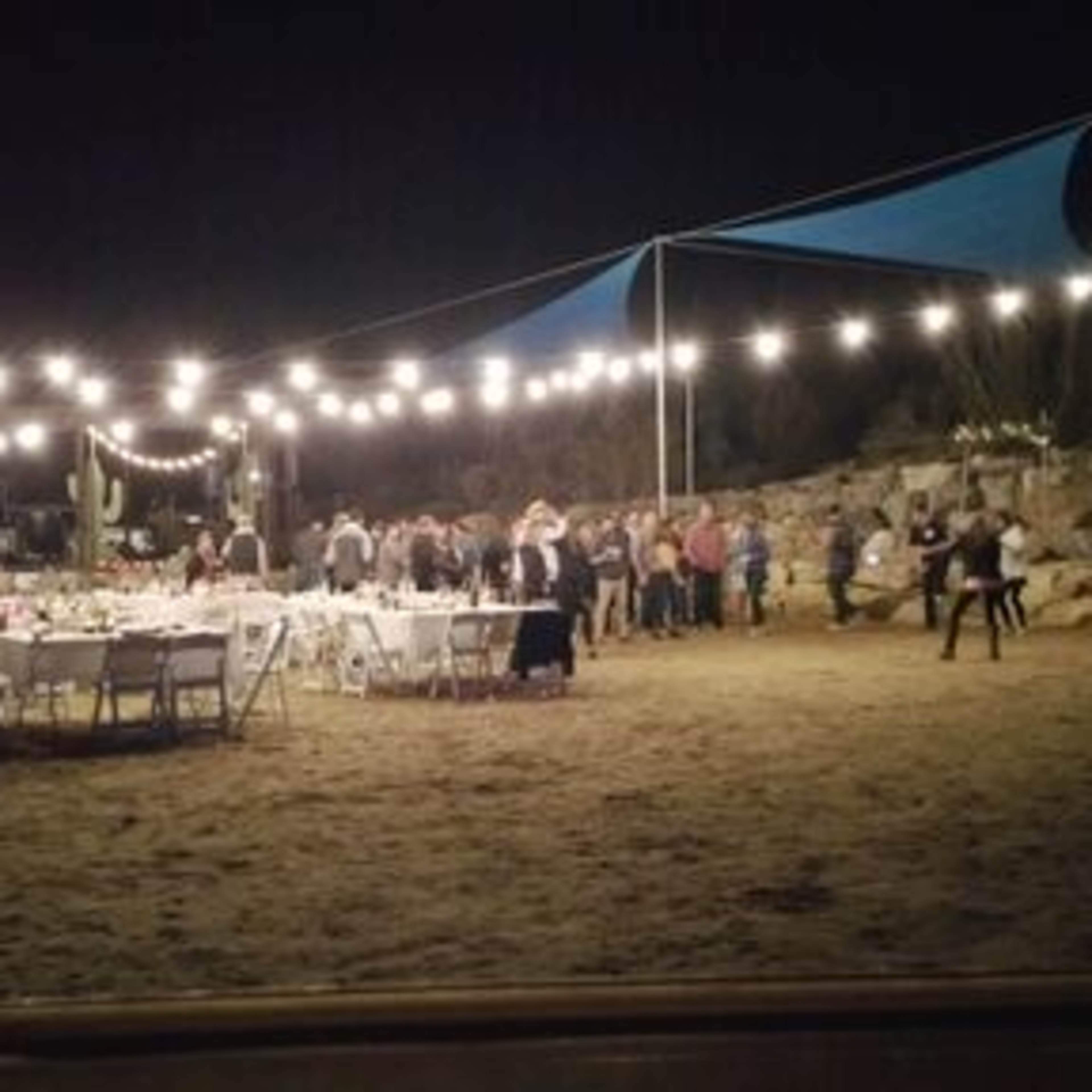 A nighttime outdoor event features tables set with decorations and string lights illuminating a gathering of people among rocky terrain.