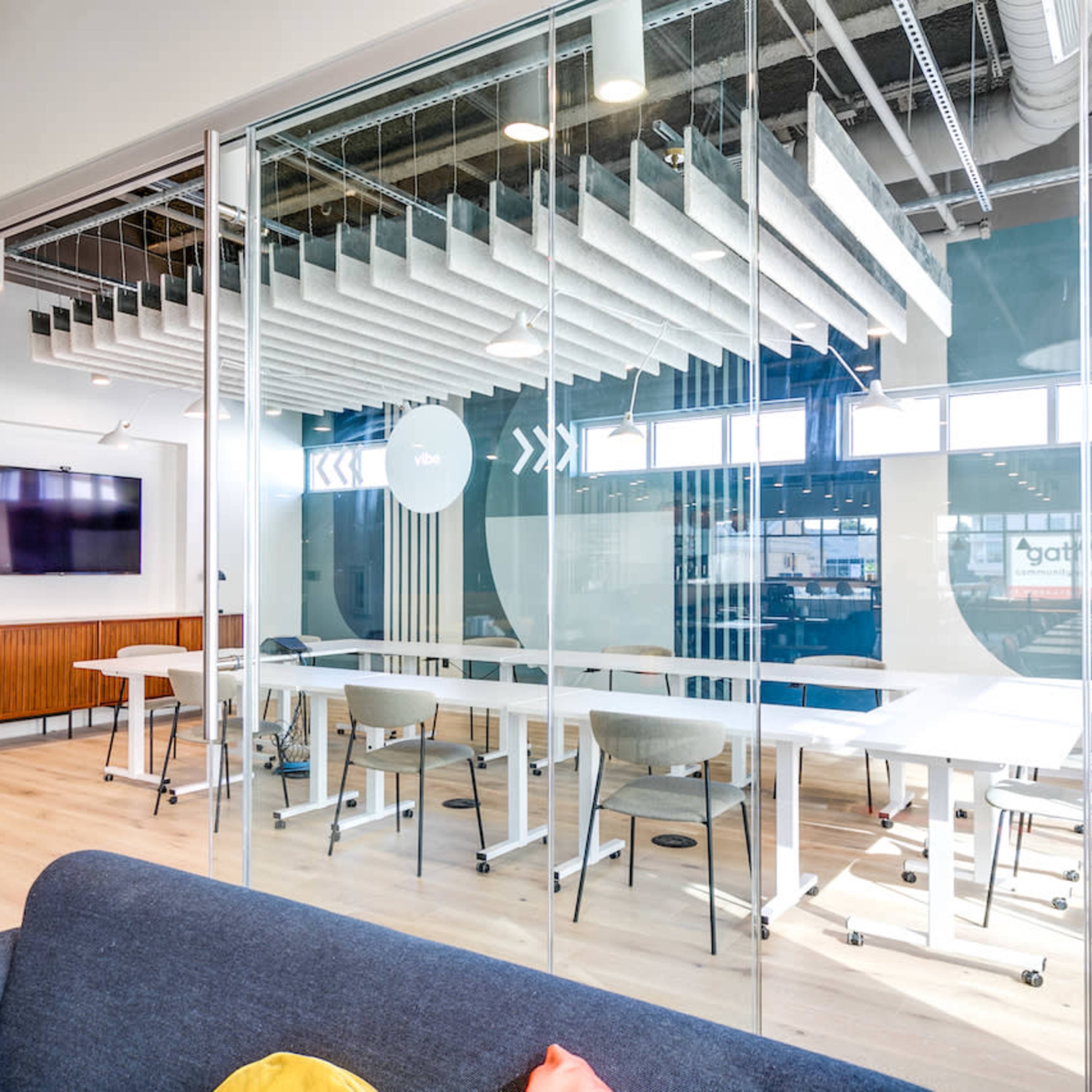 A modern office conference room features a large glass wall, white tables, and a wall-mounted television.
