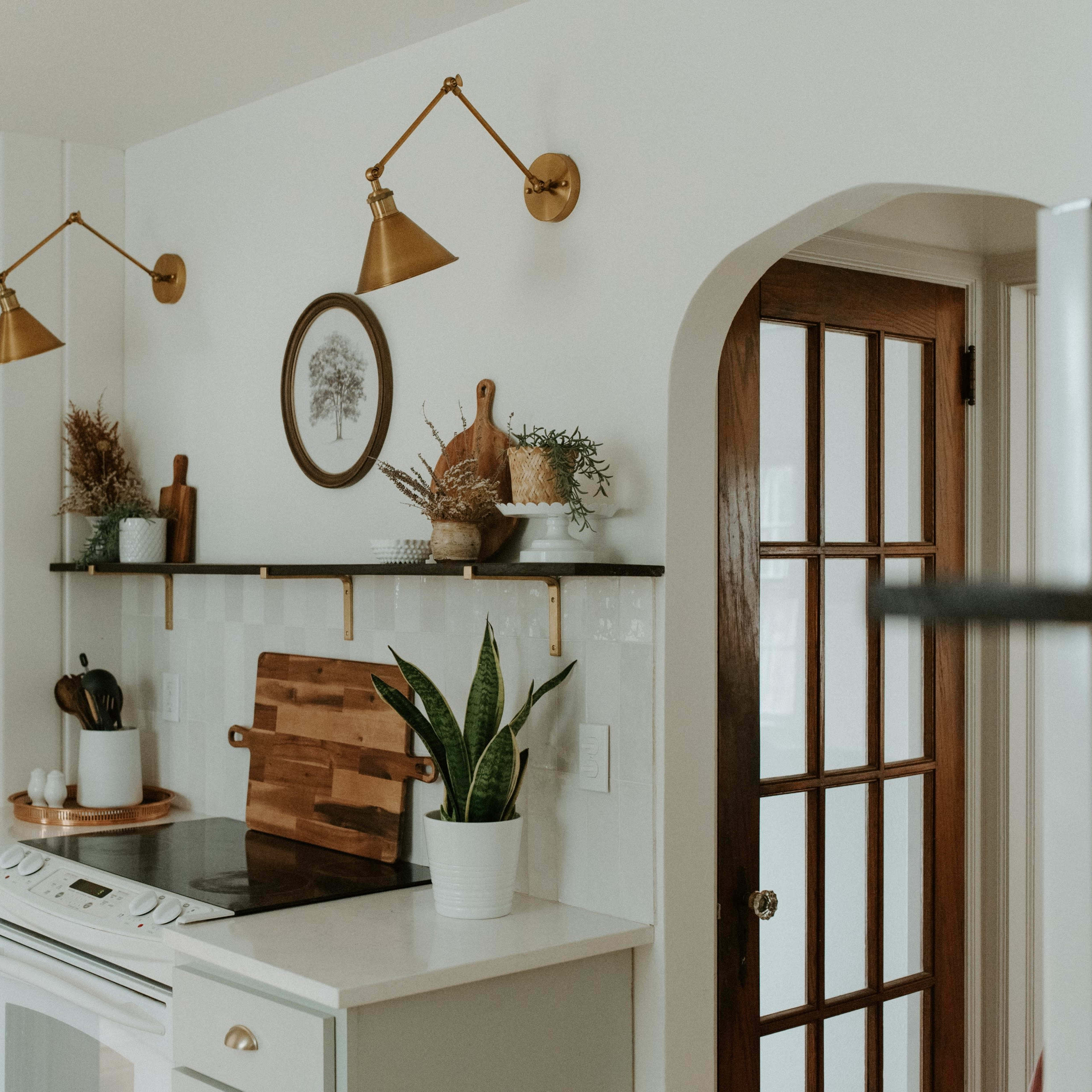 A kitchen corner with a stove, wooden cutting board, wall shelves, decorative plants, and two brass wall sconces.