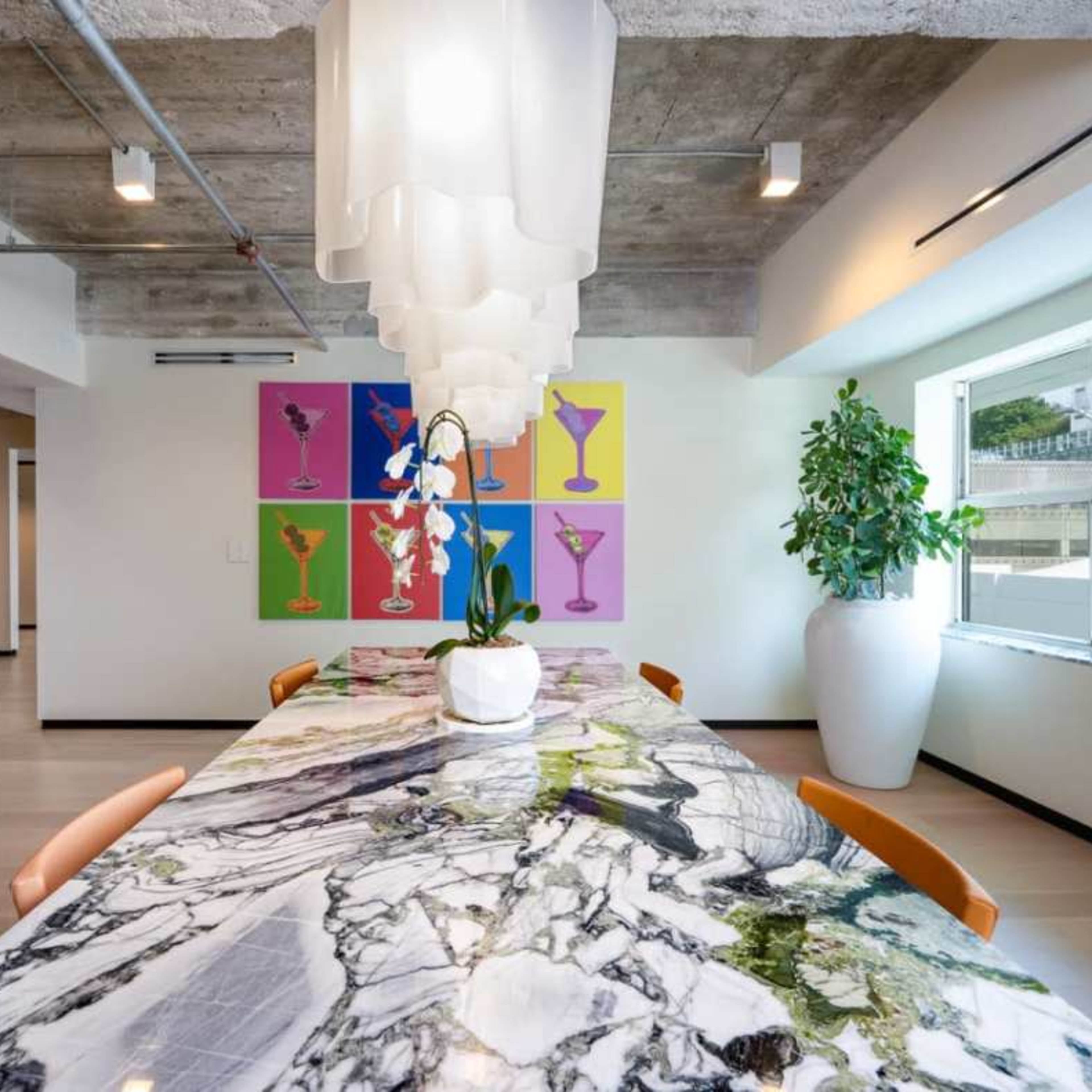A modern dining area featuring a large marble table with a centerpiece plant, surrounded by orange chairs, and a colorful art display on the wall.
