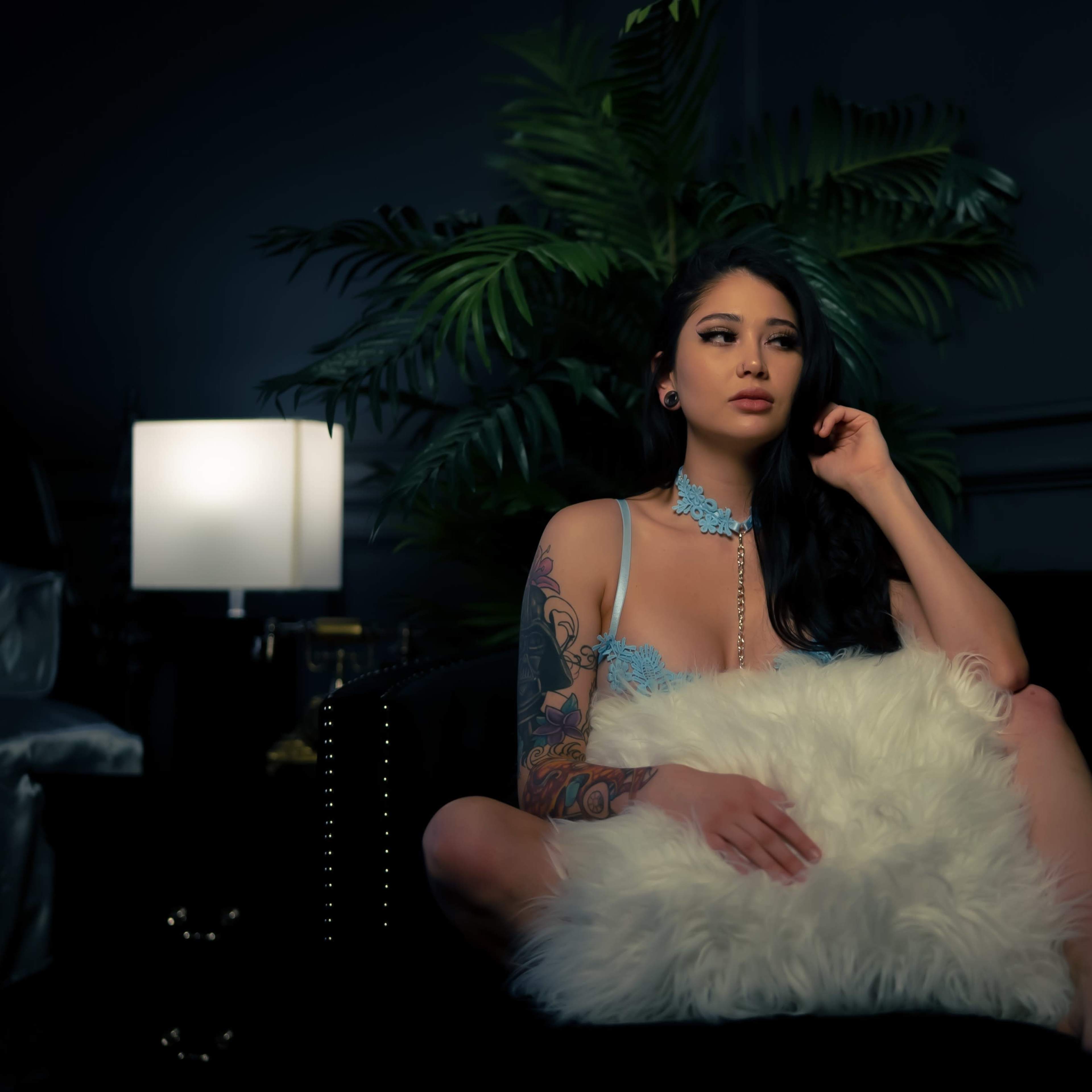 A person sits on a sofa holding a fluffy pillow, surrounded by indoor plants and soft lighting.