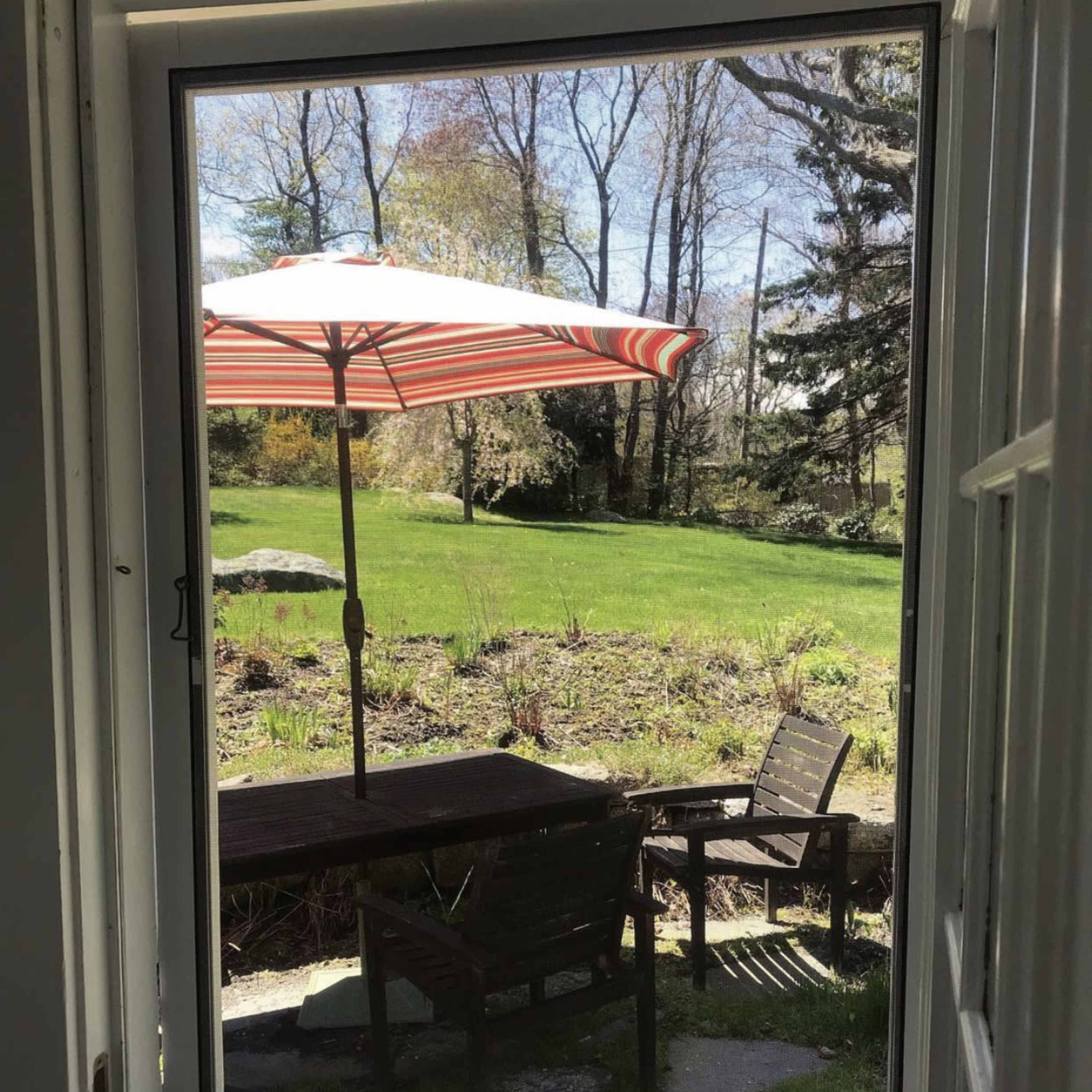A view through an open door reveals a patio area with a striped umbrella over a table surrounded by chairs, set against a green lawn and trees in the background.