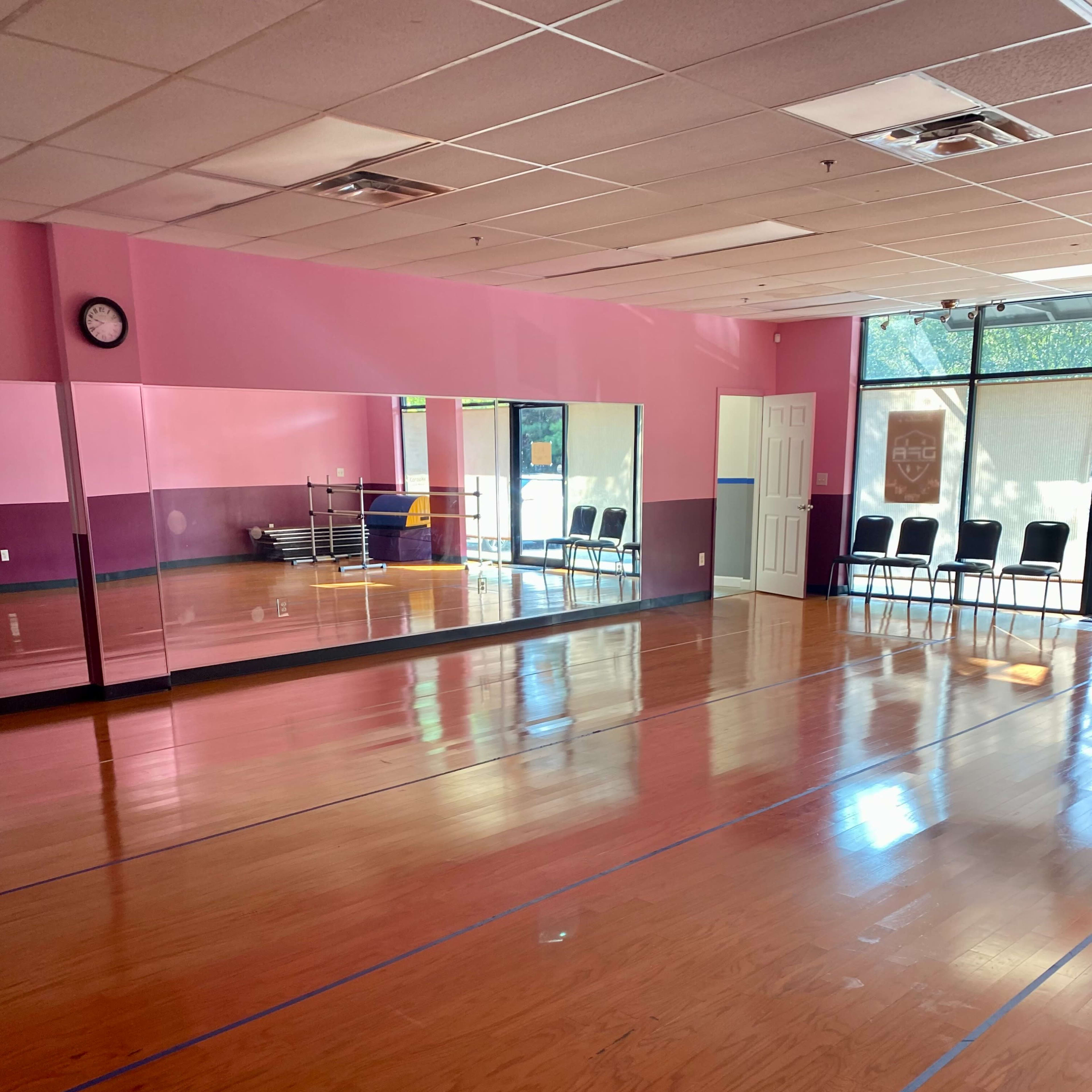 A spacious dance studio with hardwood floors, pink walls, large mirrors, and a set of chairs lined against the wall.