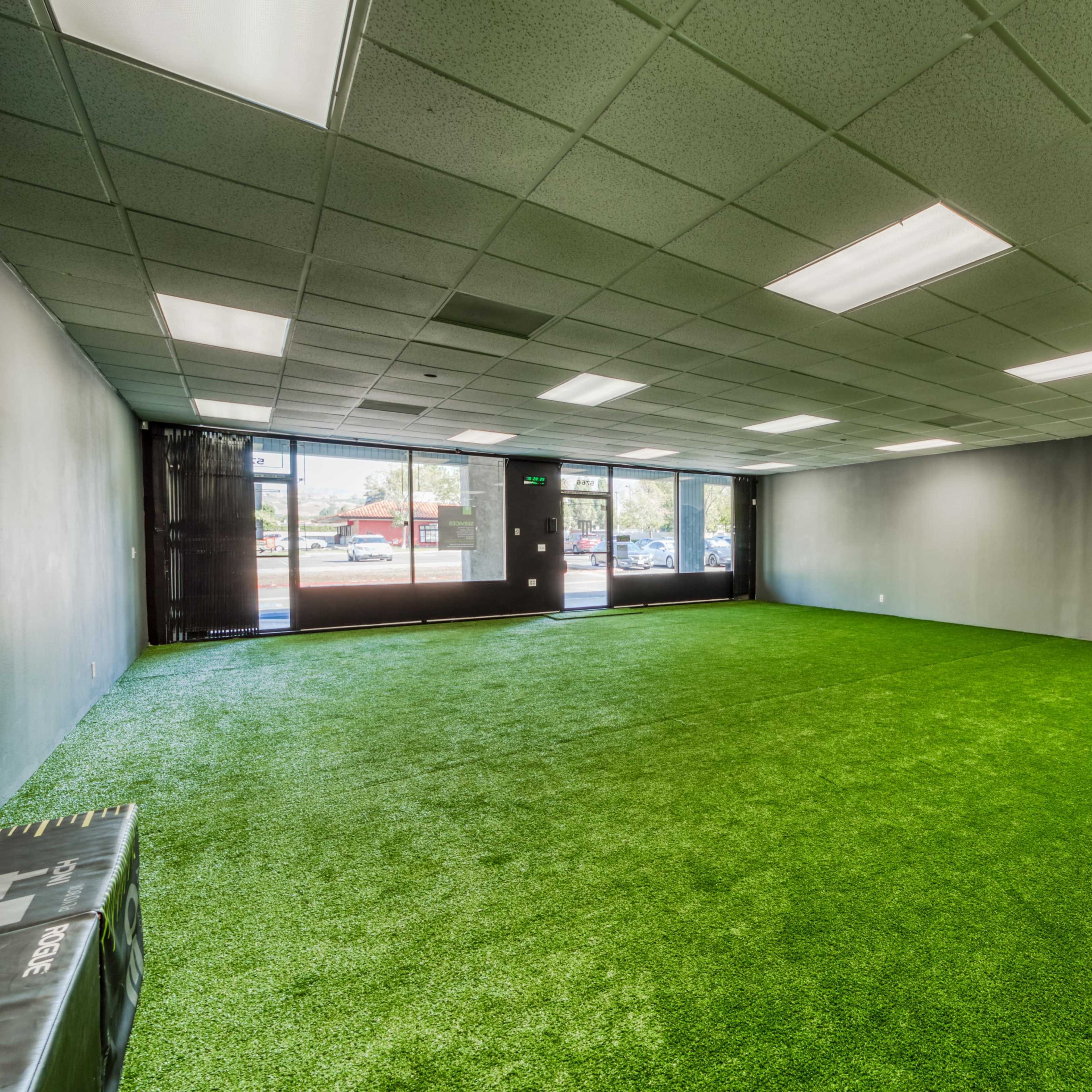 An empty room with artificial grass flooring, large windows allowing natural light, and fitness equipment positioned in one corner.