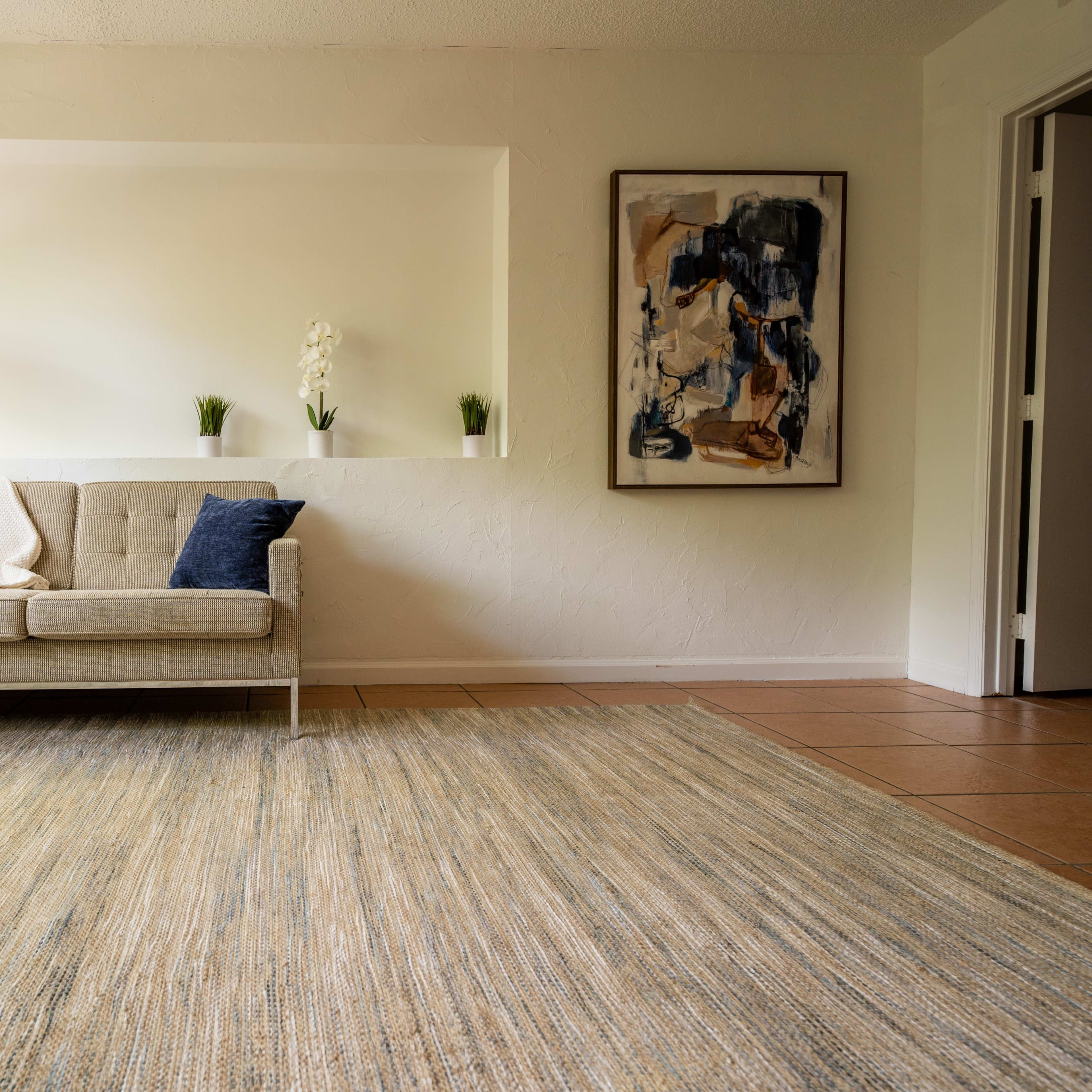 A simple living room featuring a light-colored sofa with blue pillows, a textured area rug, and a wall adorned with a framed artwork.
