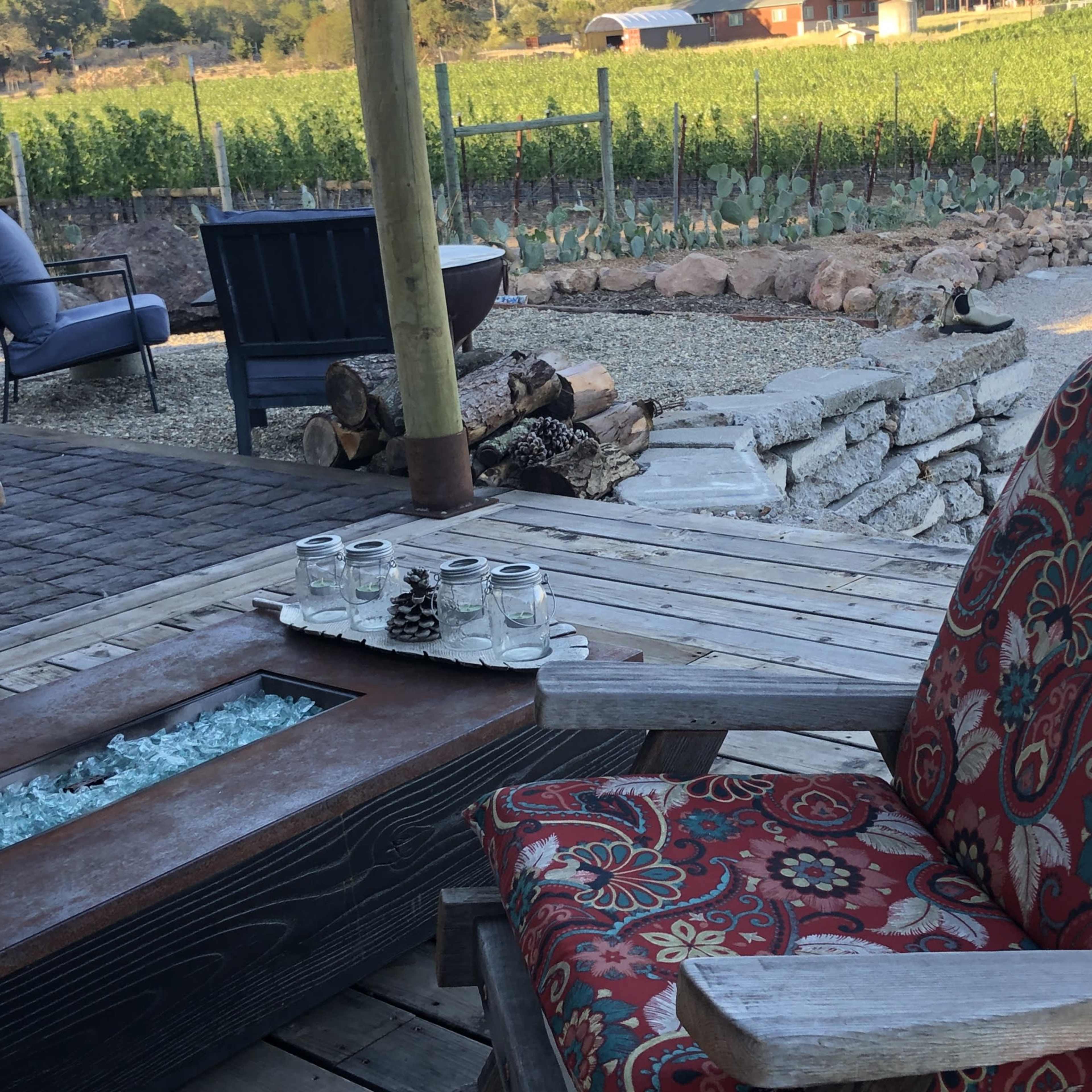 A cozy outdoor seating area with patterned chairs surrounding a fire pit, overlooking a vineyard in the background.