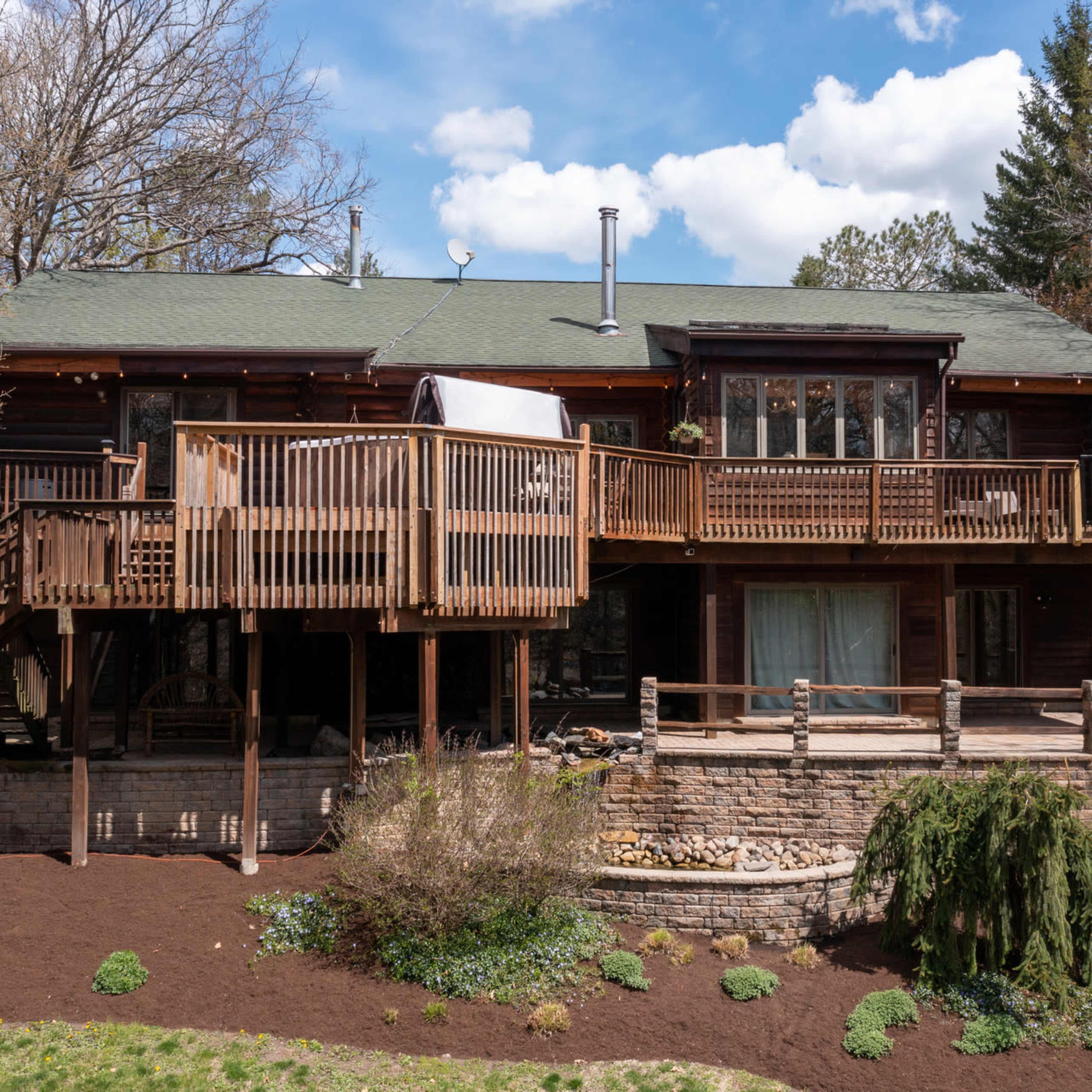 A wooden two-story house with a large deck supported by wooden posts, surrounded by trees and landscaping.