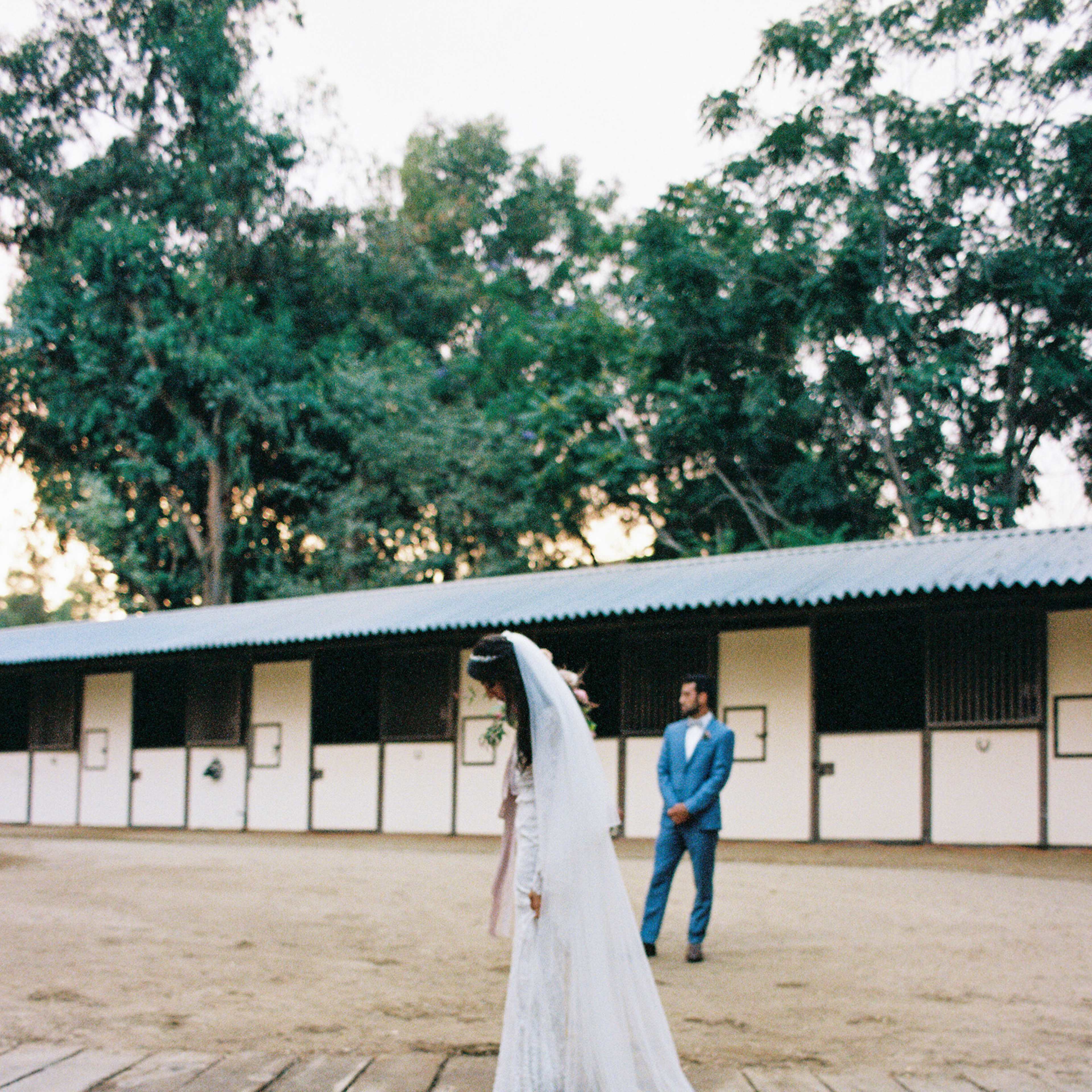 A bride in a white dress and veil stands on a wooden pathway, while a man in a blue suit watches from a distance at an outdoor venue.
