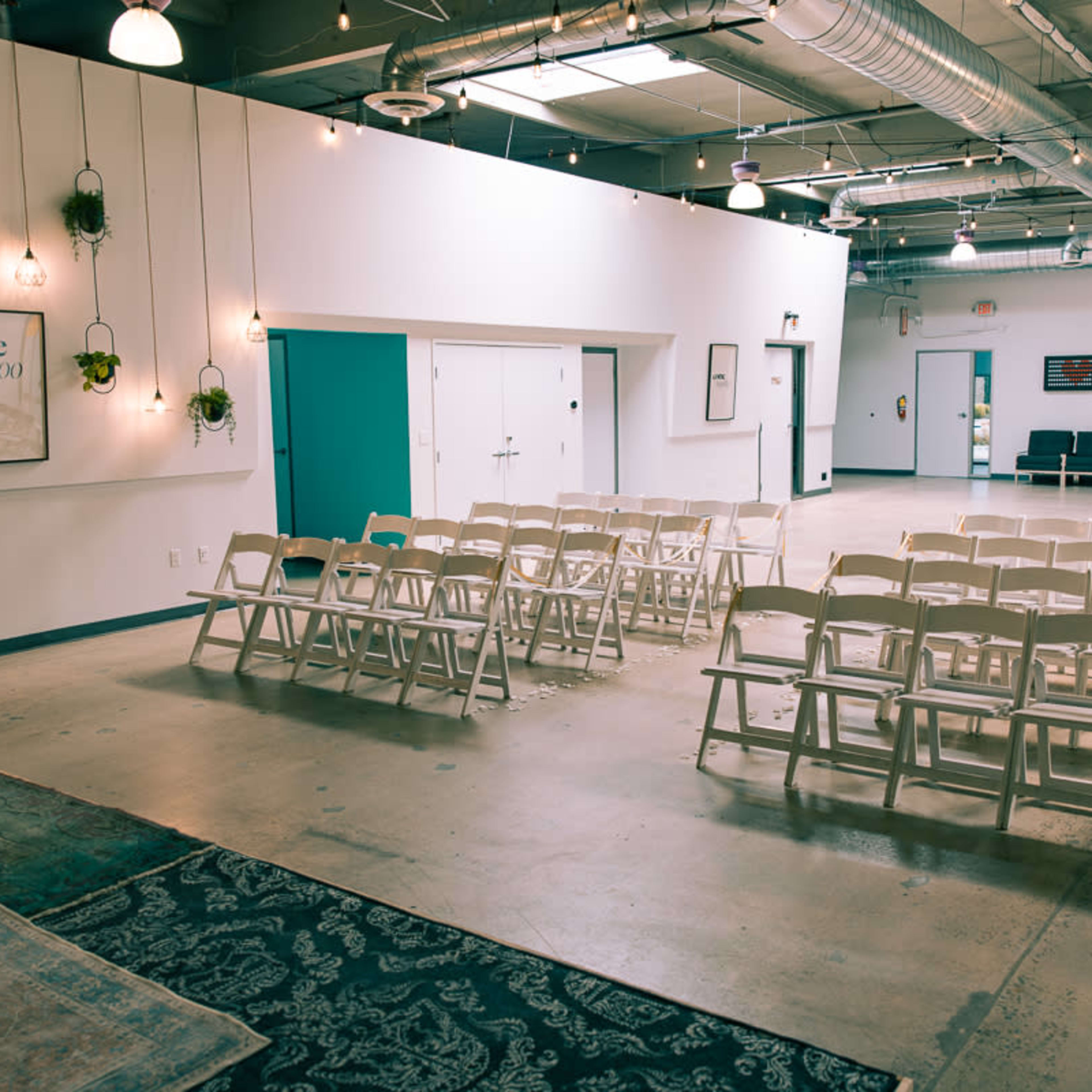 A spacious room set up for an event, featuring rows of white chairs arranged in front of a wall adorned with decorative plants and framed artwork.