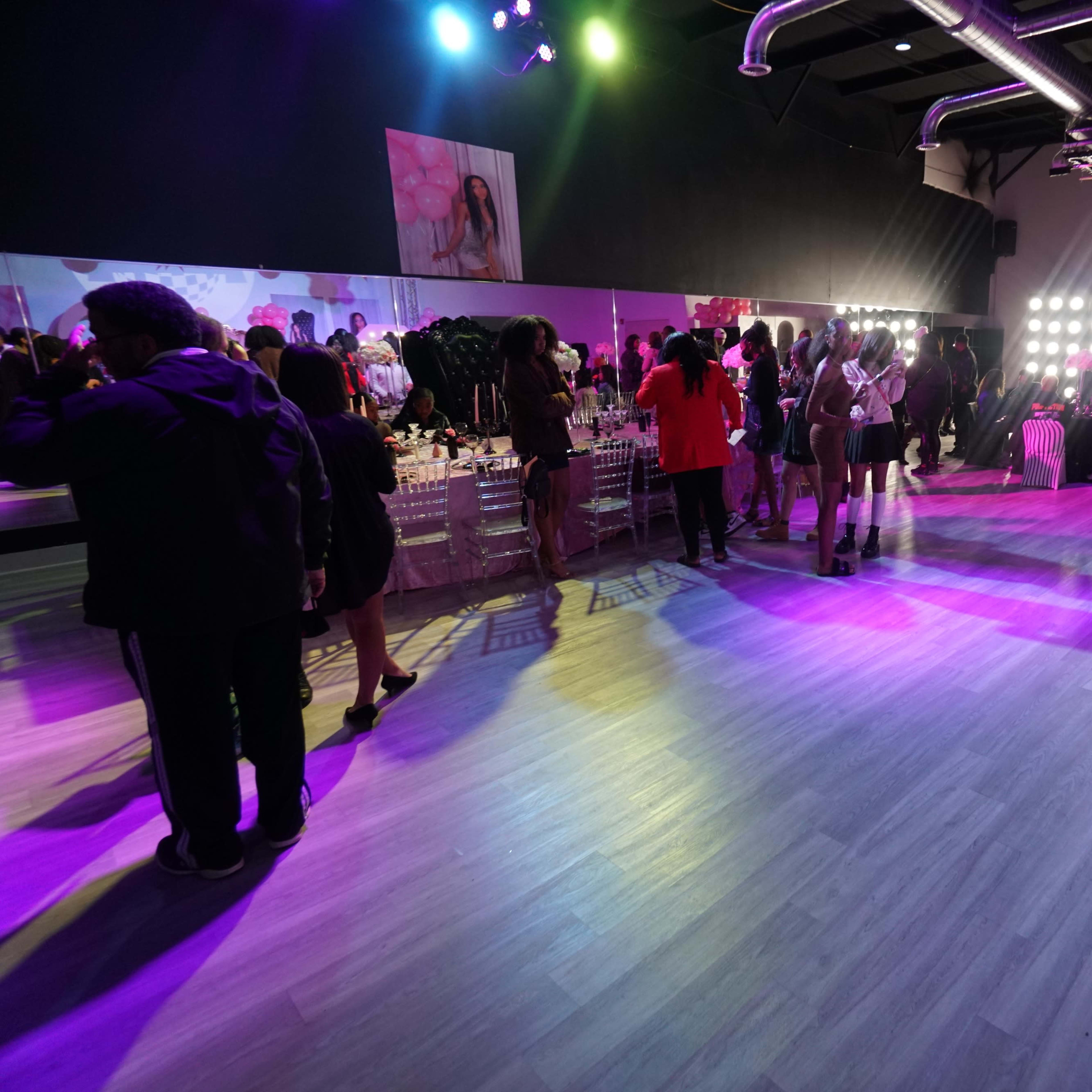 A crowded event space is filled with people socializing under colorful lighting, with tables set up for a celebration on one side.