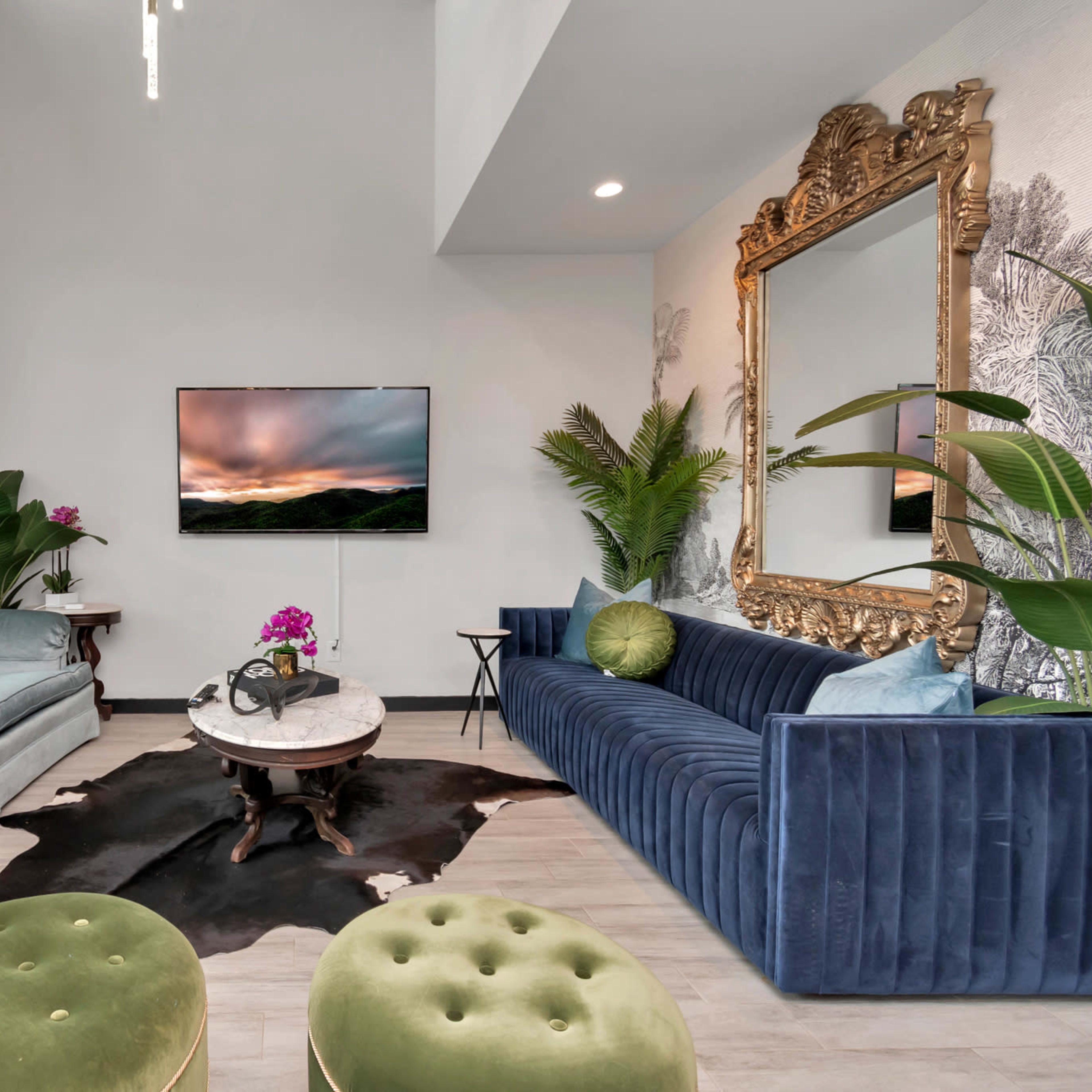A stylish living room featuring a blue velvet sofa, a gray couch, a round coffee table, green plants, and a large golden mirror above the television.