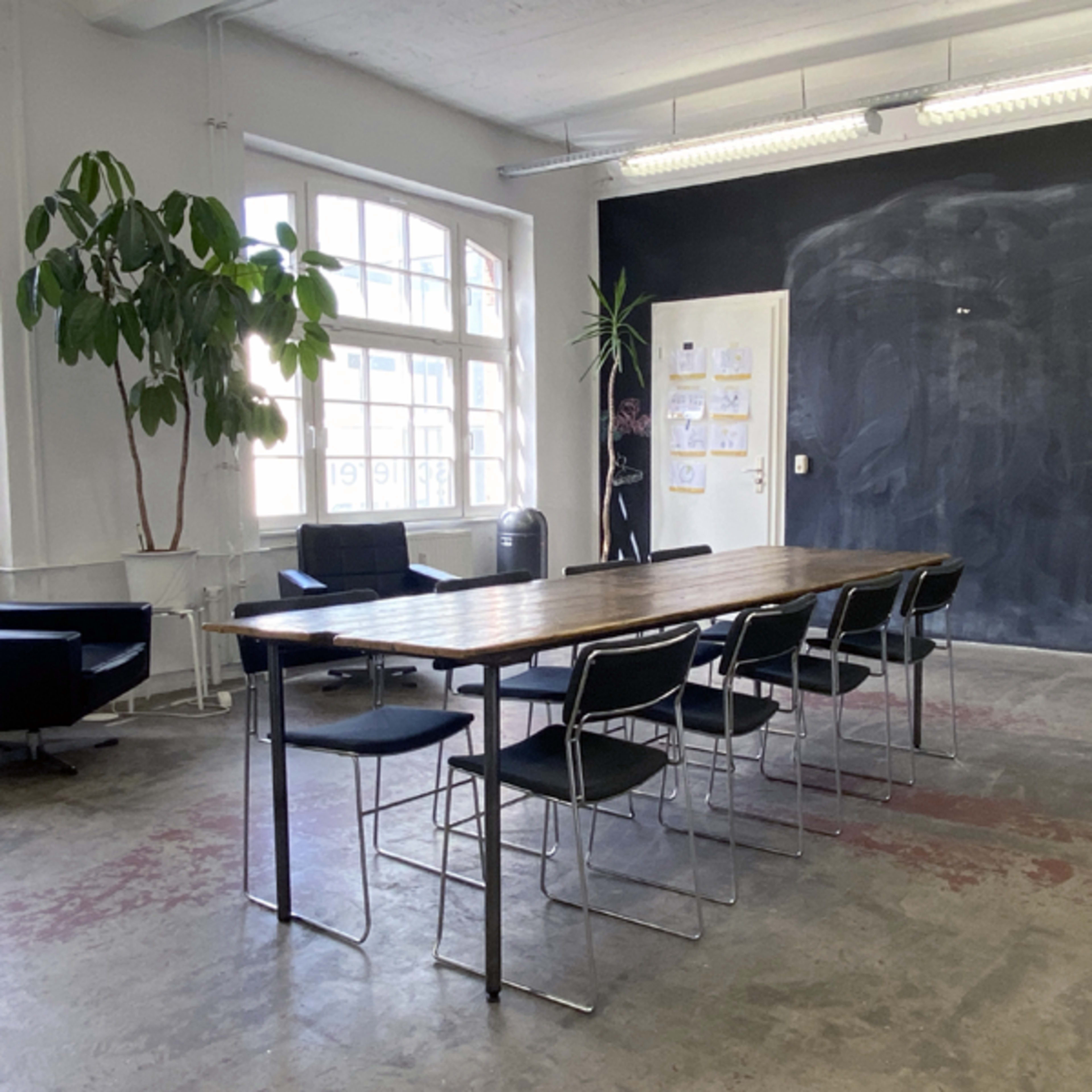 A spacious meeting room featuring a long wooden table surrounded by black chairs, with large windows and a chalkboard wall.