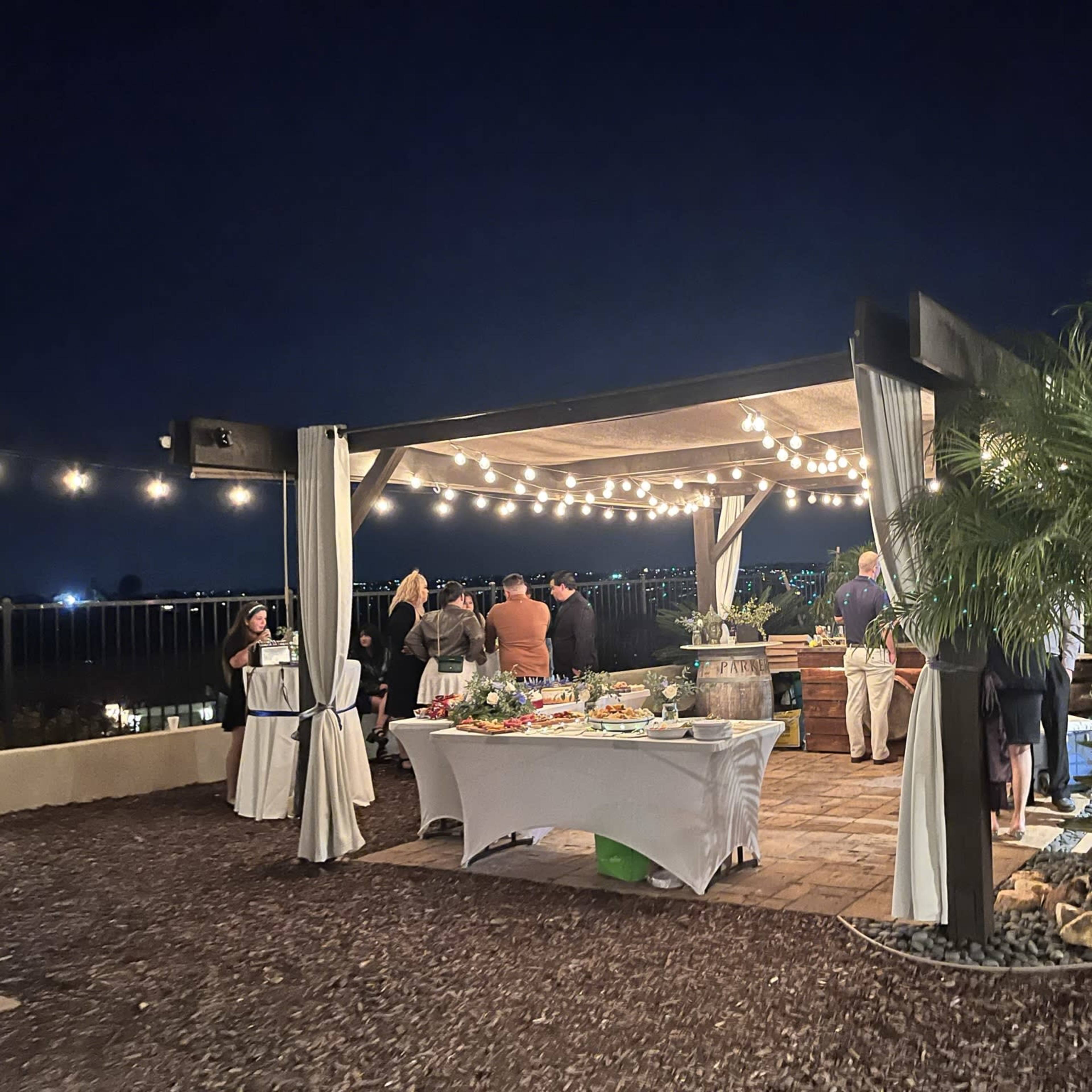 A gathering of people is having an evening event under a beautifully lit pavilion with tables featuring food and drinks, surrounded by palm trees and city views.