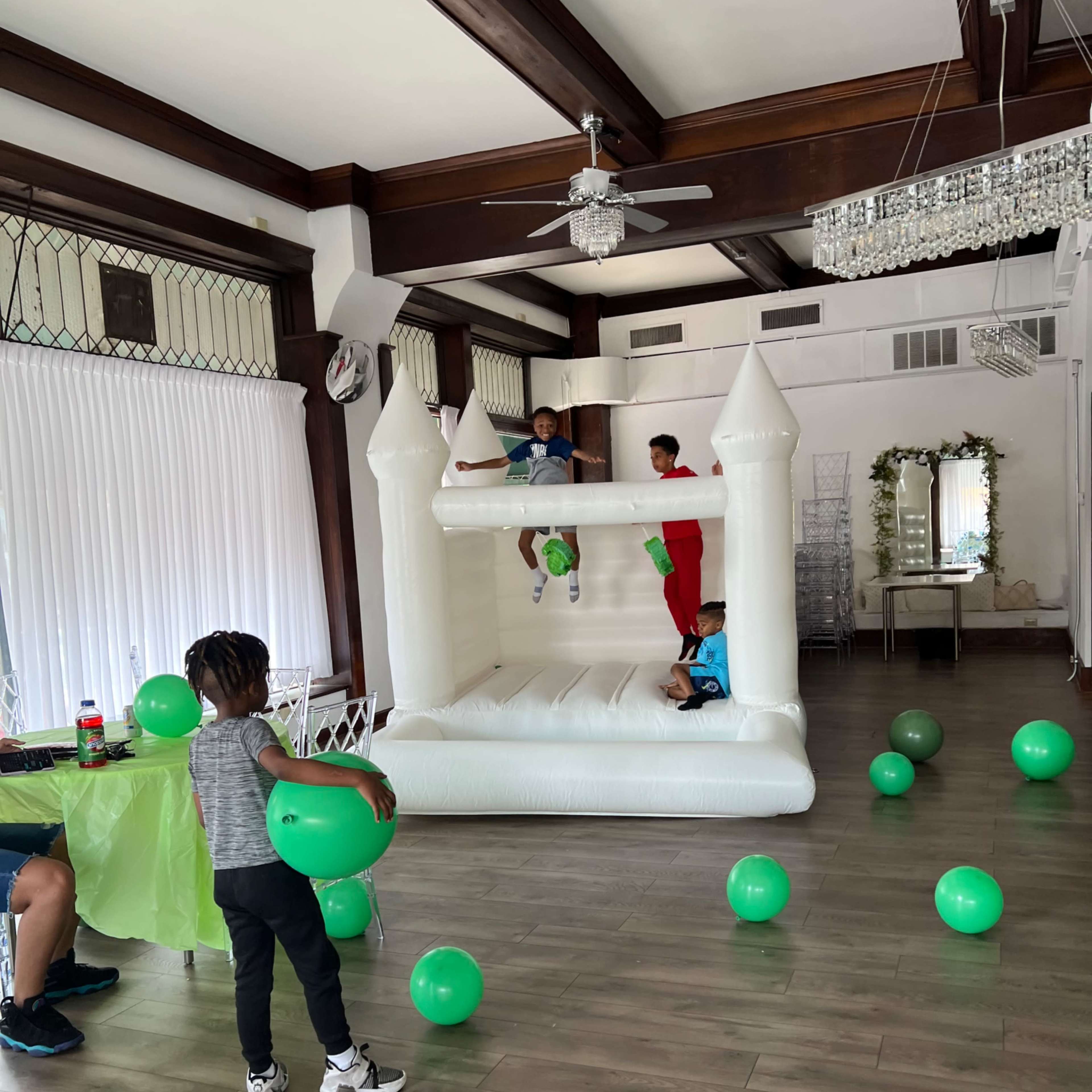 Children are playing in a white bounce house while others hold green balloons in a brightly lit event space.