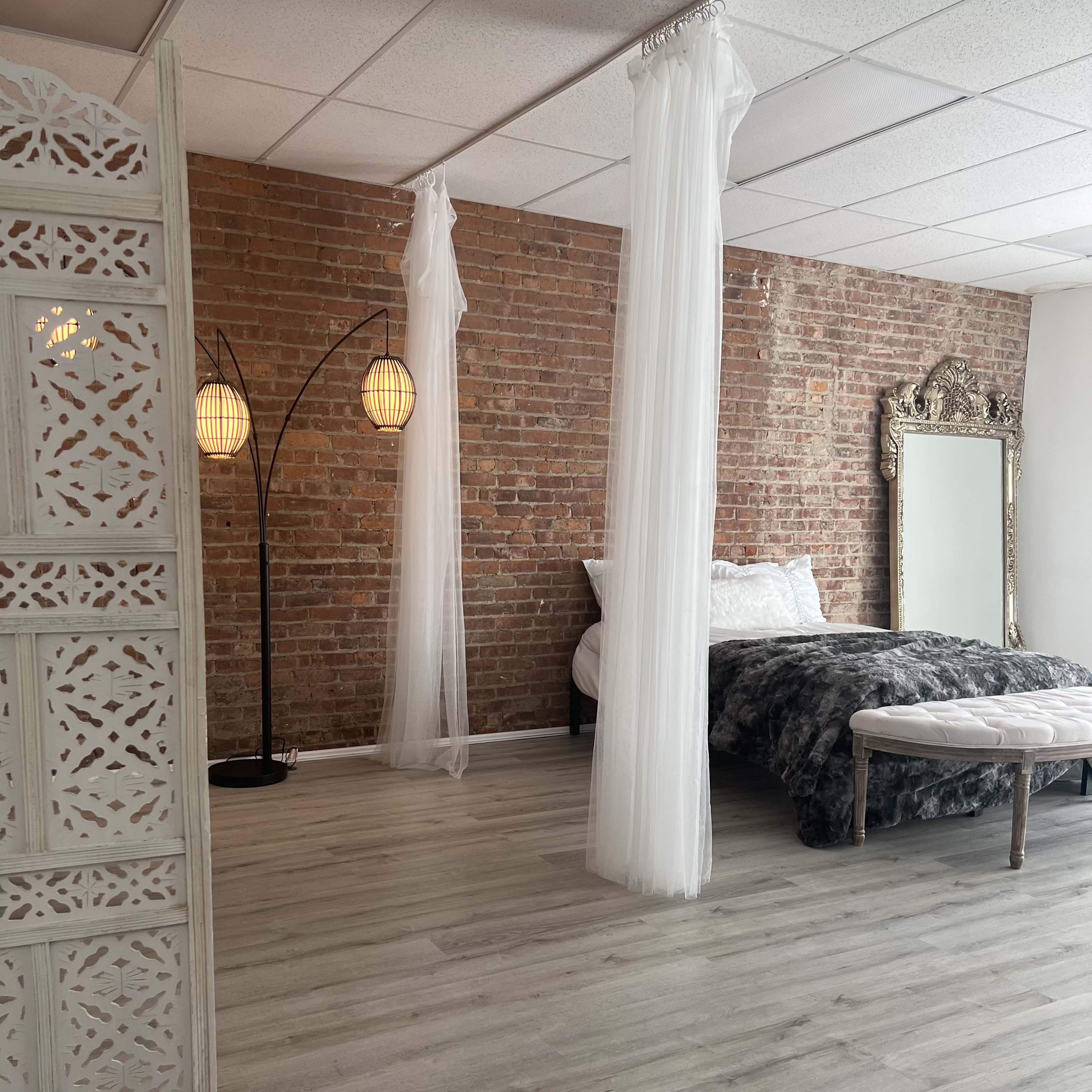 A bedroom with exposed brick walls, a bed draped in gray bedding, sheer white curtains hanging from the ceiling, and a decorative mirror beside a modern floor lamp.