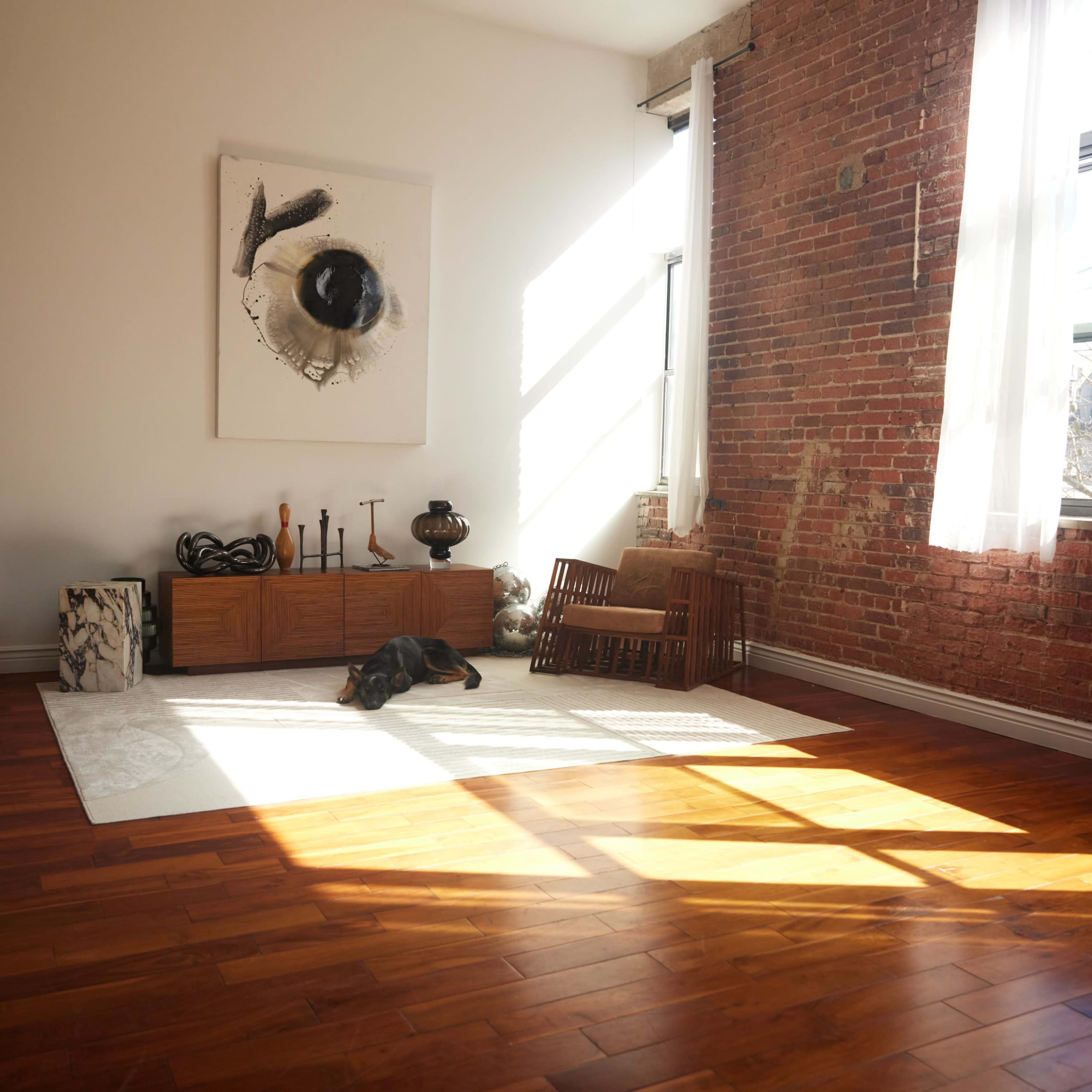 A spacious room features exposed brick walls, wooden flooring, and a large window casting shadows on a white area rug.