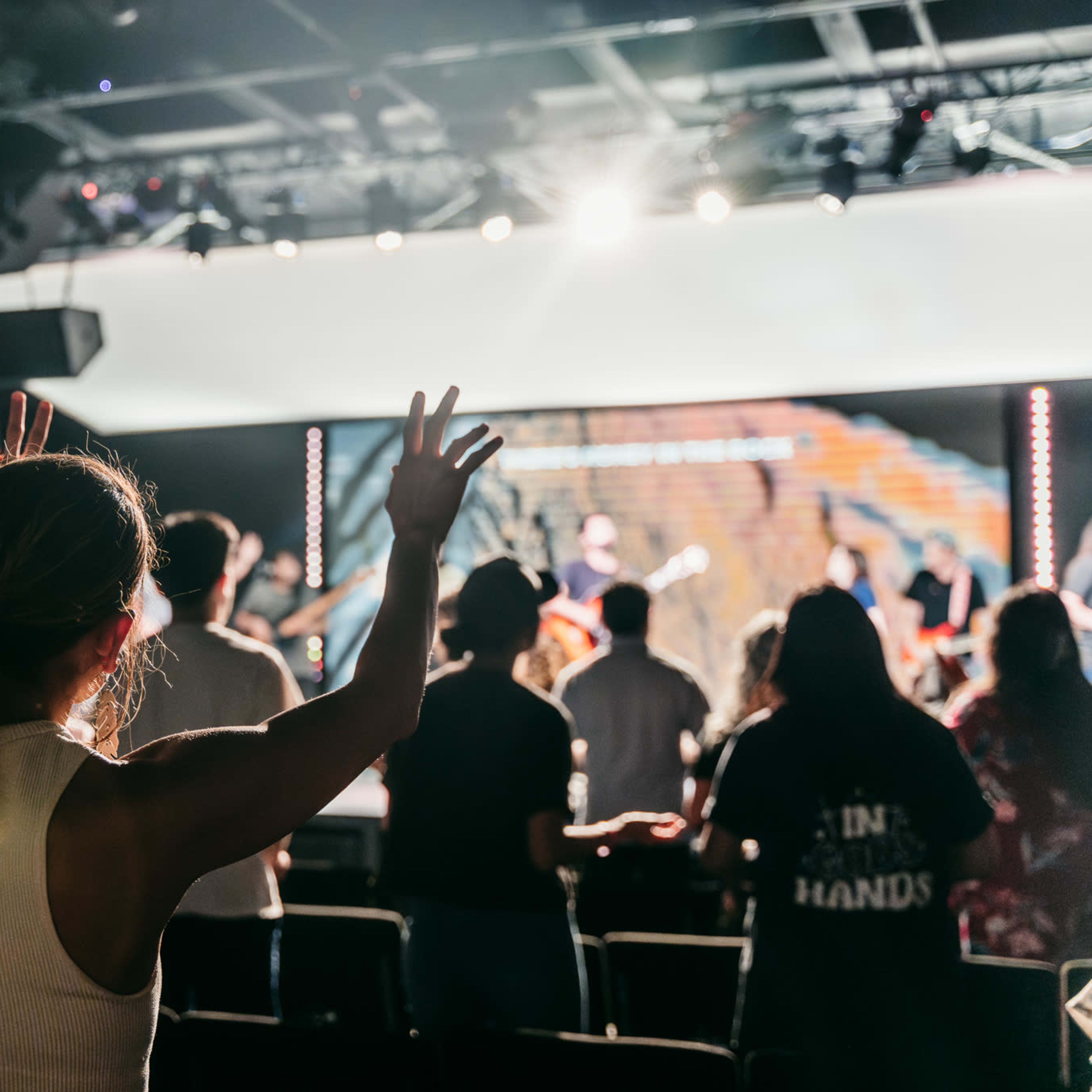 A group of people with raised hands worship during a live music performance in a dimly lit venue.