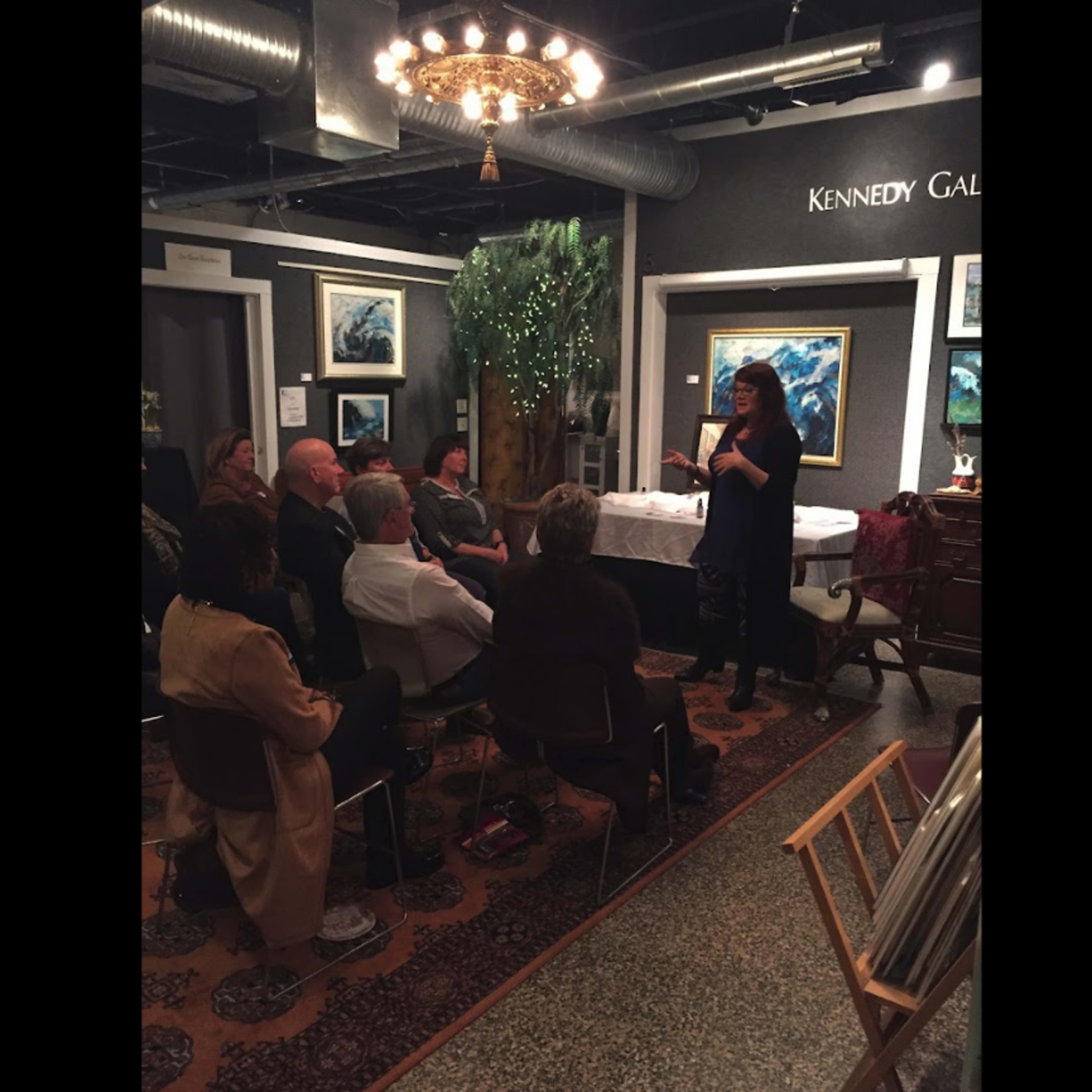 A speaker presents to an audience seated in a gallery adorned with artwork and plants.