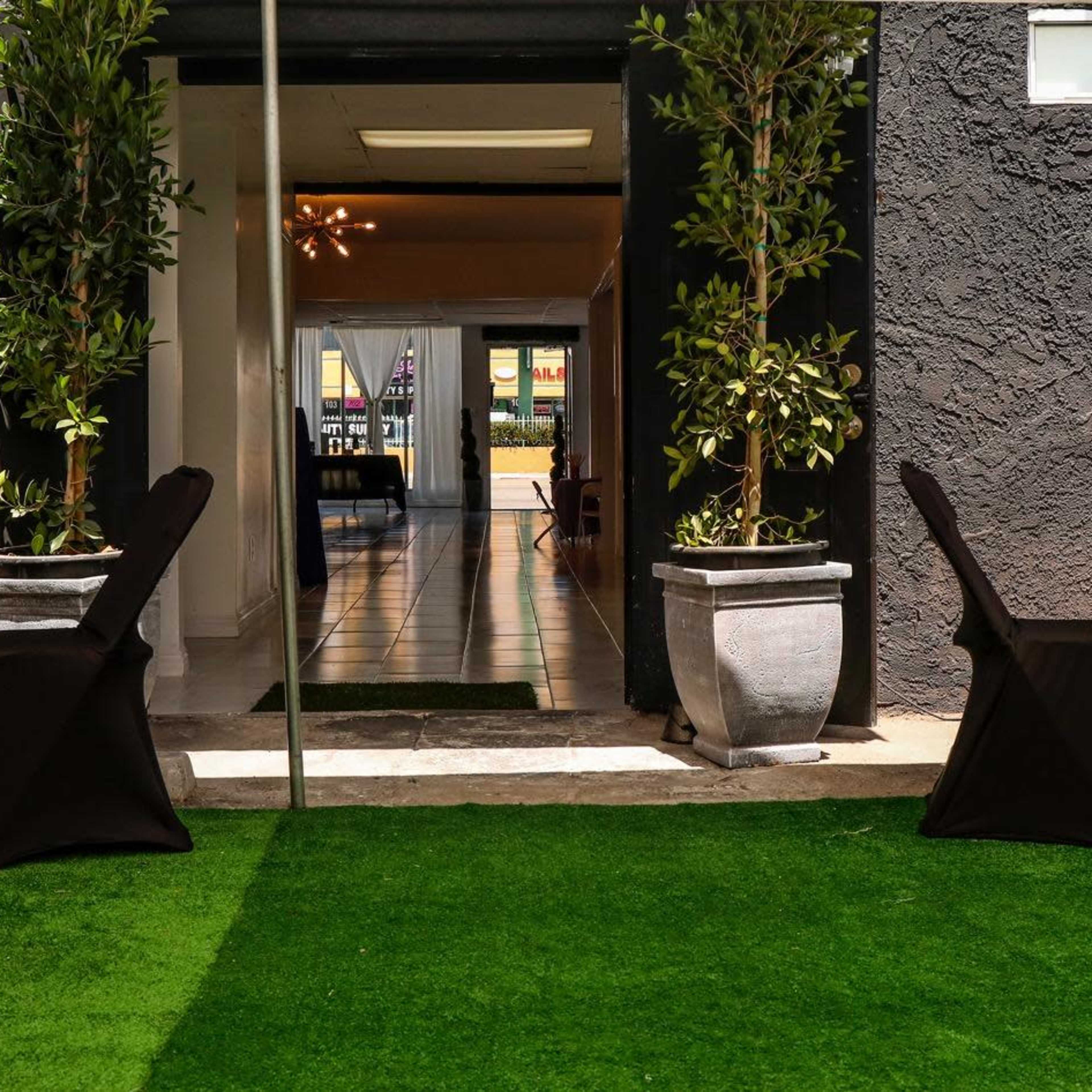 An outdoor event space with black chairs and tables set on artificial grass, leading to a doorway into a brightly lit interior.