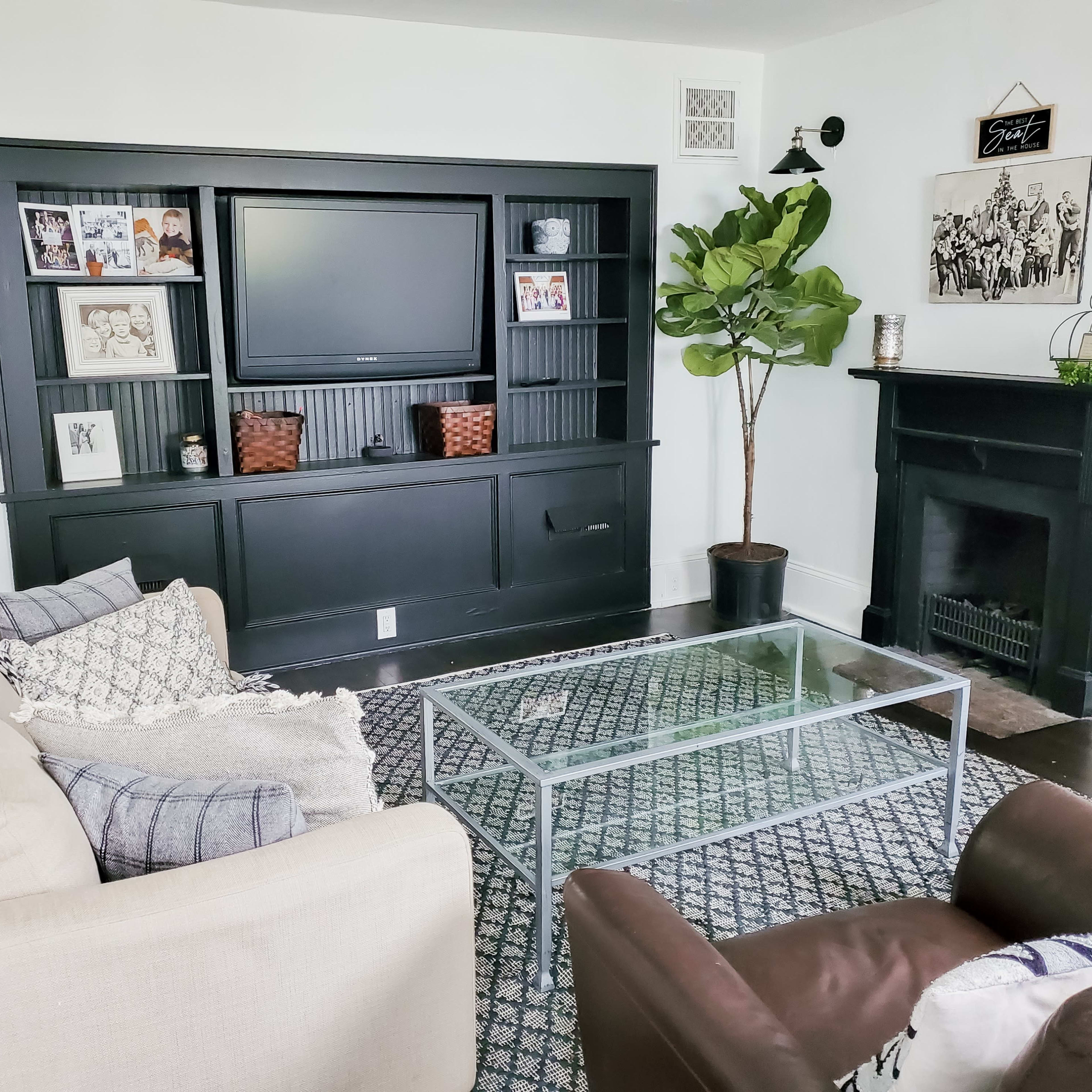 A living room with a black entertainment unit, a glass coffee table, a light-colored sofa, two brown armchairs, and a fireplace.